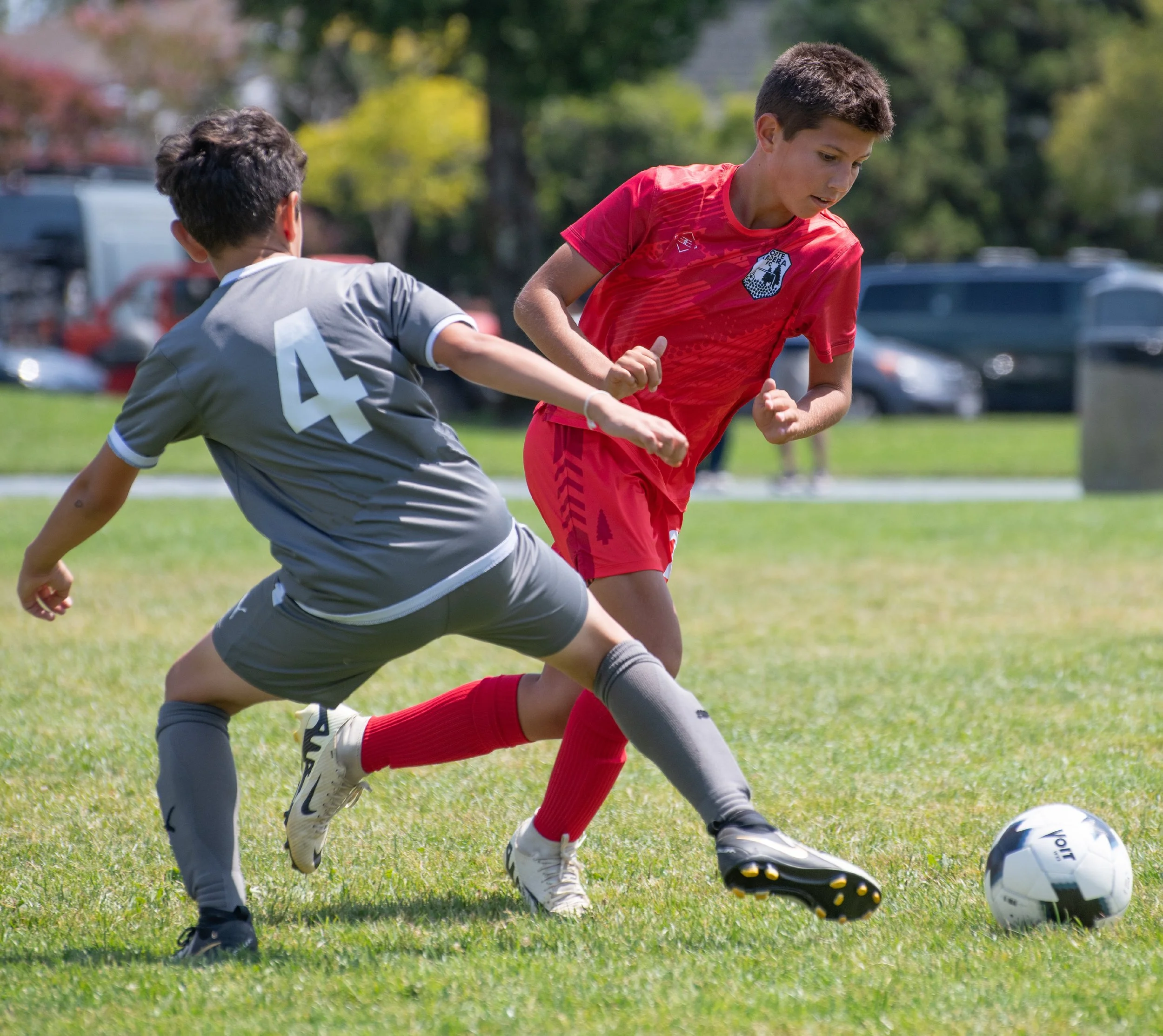 Two young boys playing soccer outdoors on grass, one in a gray jersey with the number 4, and the other in a red jersey, both trying to gain control of a soccer ball.
