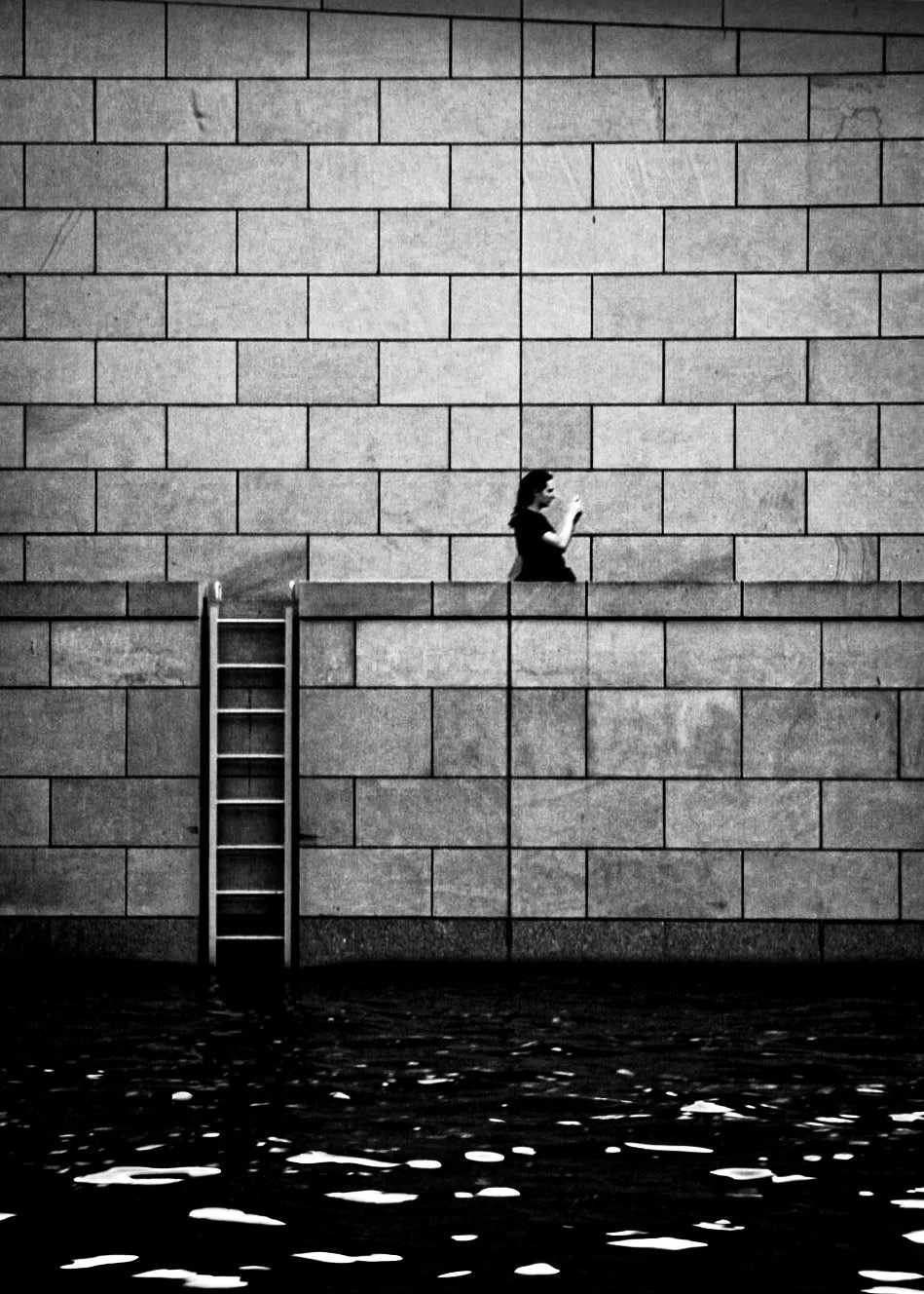 This photograph feels like a quiet, cinematic moment suspended between worlds.

In the frame, a person walks along a high stone wall, their silhouette stark against the light-toned blocks. The wall itself dominates the image — its geometric pattern o