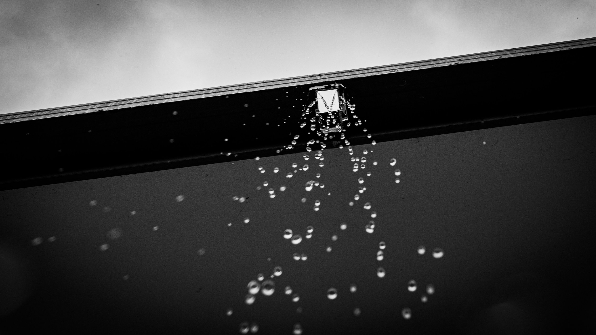 This photograph captures a fleeting, contemplative moment, rendered in stark black and white. From the edge of a roof above, a thin stream of water breaks apart into individual droplets, each one suspended briefly as it falls. The droplets hang in th