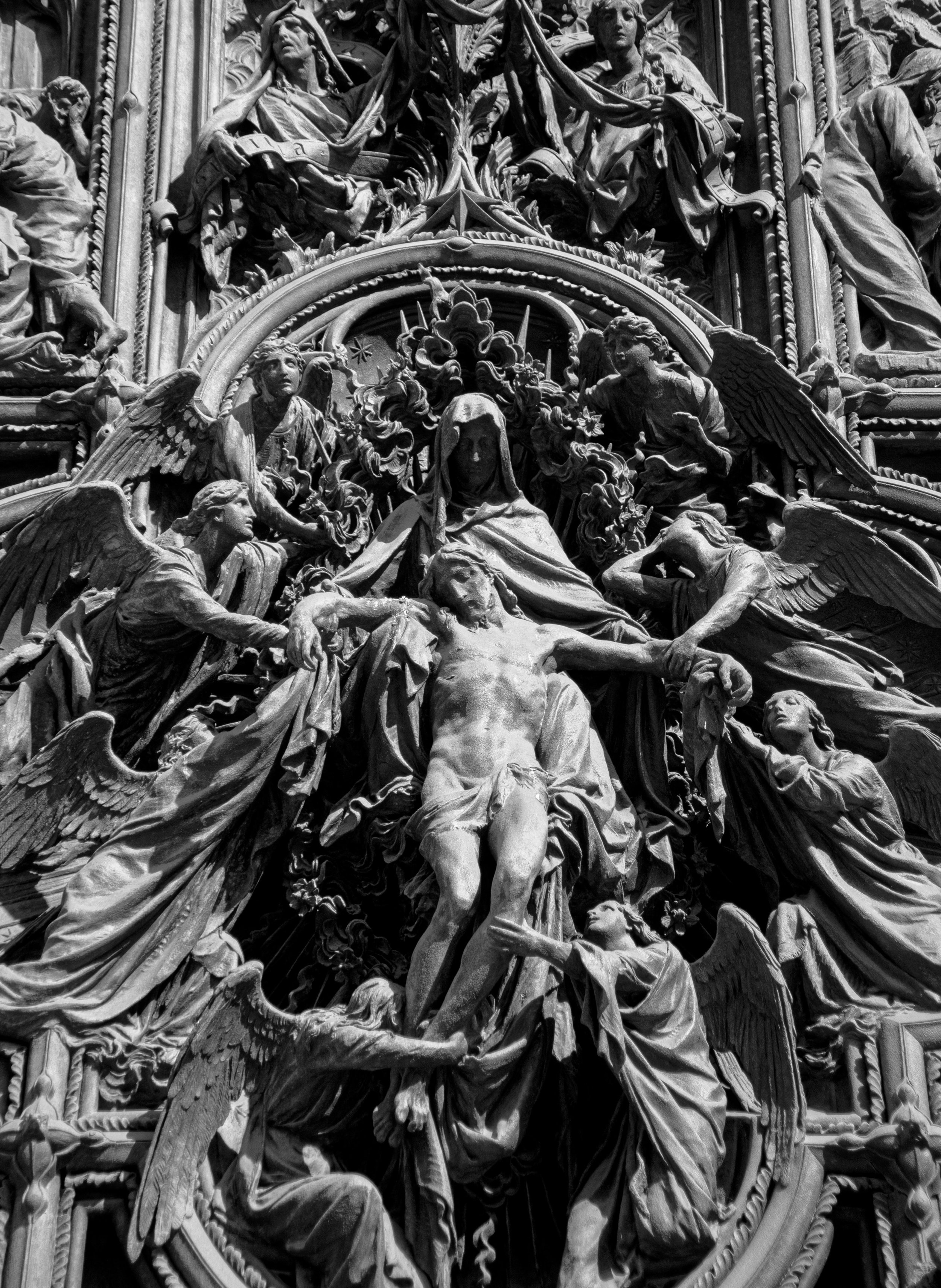 This photograph reveals a sculptural scene that feels less like carved stone and more like the crystallization of a moment suspended between worlds — the point where human sorrow meets divine embrace. The monochrome palette strips away distraction, l