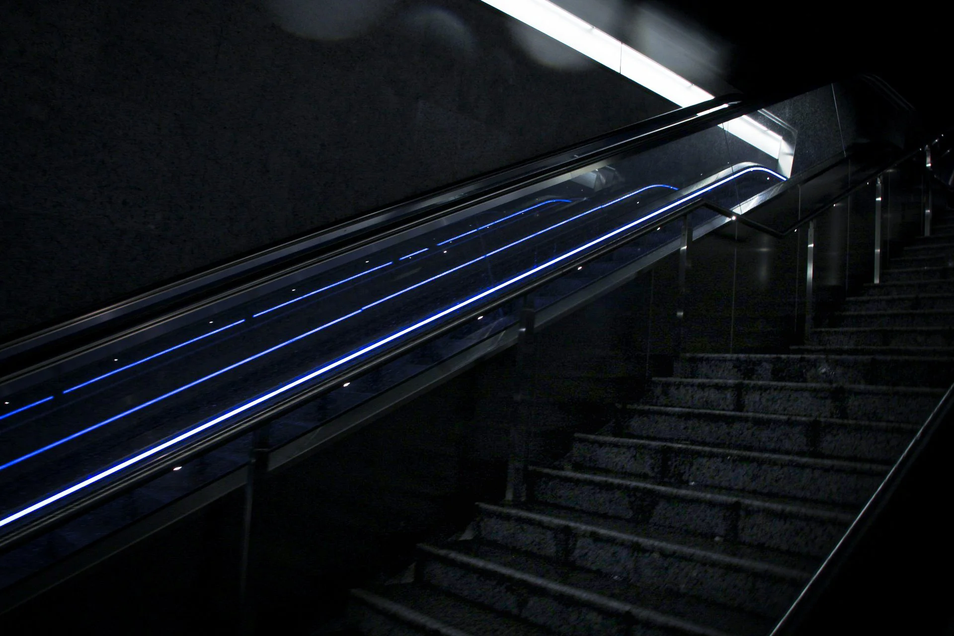 Dark interior stairwell with an illuminated escalator featuring blue LED lights along its sides.