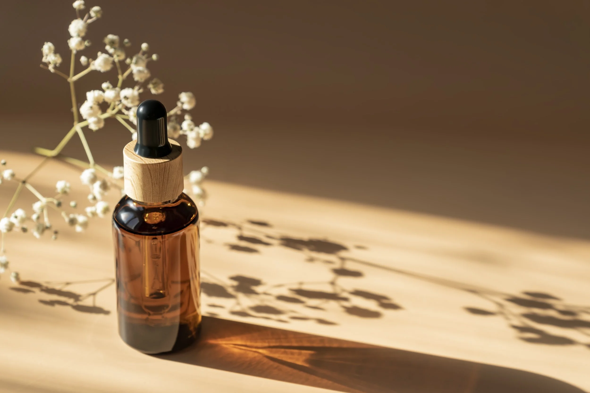 Amber glass dropper bottle on a wooden surface with white flowers and shadows.