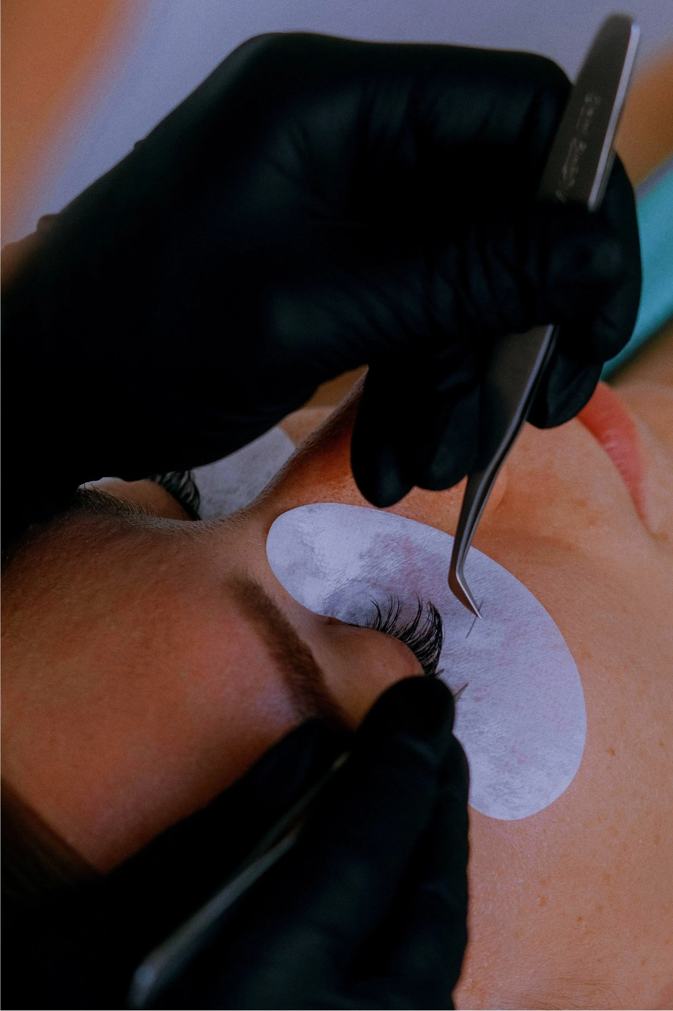 A person receiving eyelash extensions from a professional wearing black gloves, using tweezers to apply lashes on a client with an under-eye pad.