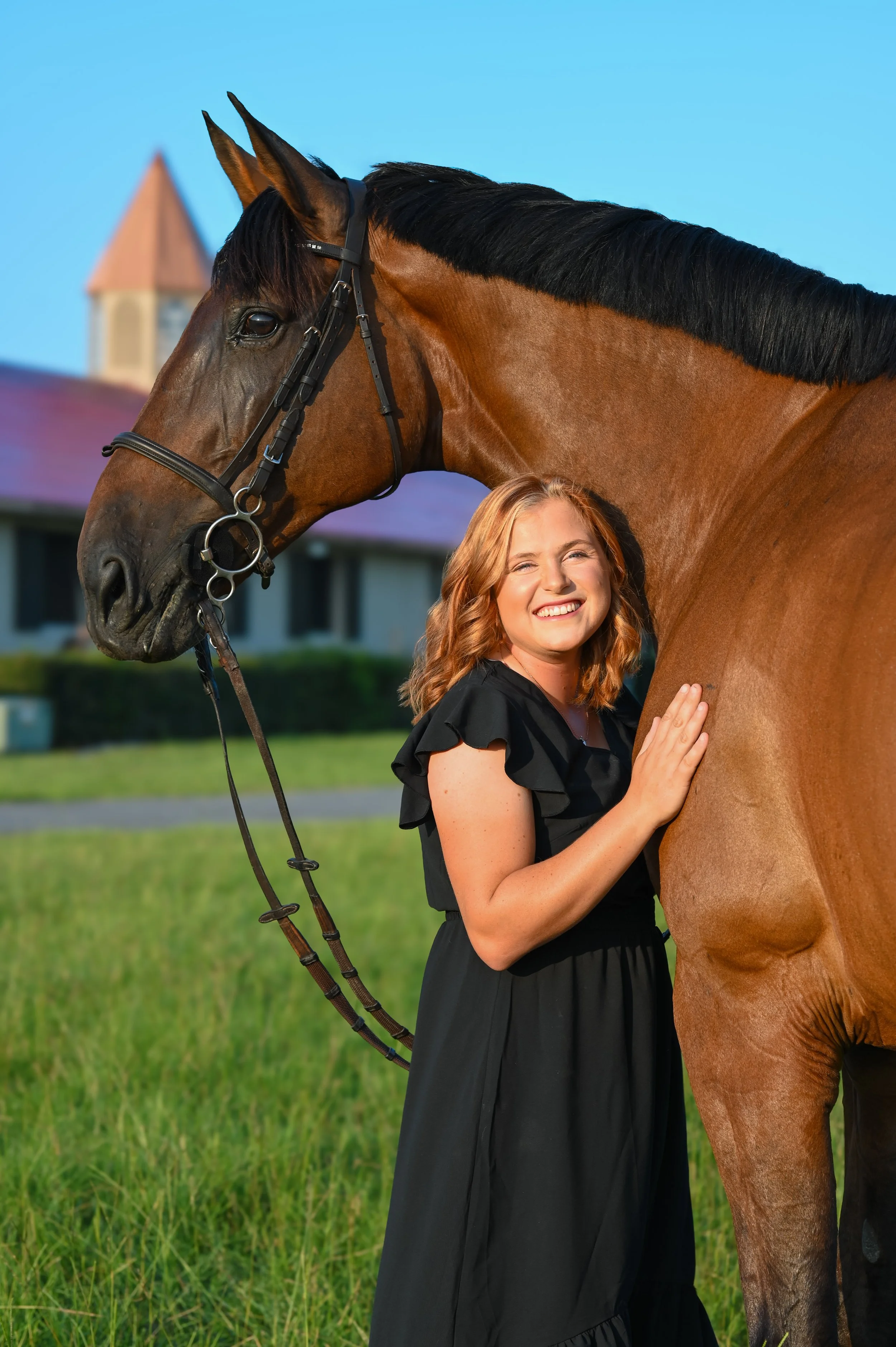 A woman with auburn hair in a black dress smiling and hugging a brown horse outdoors on a sunny day.