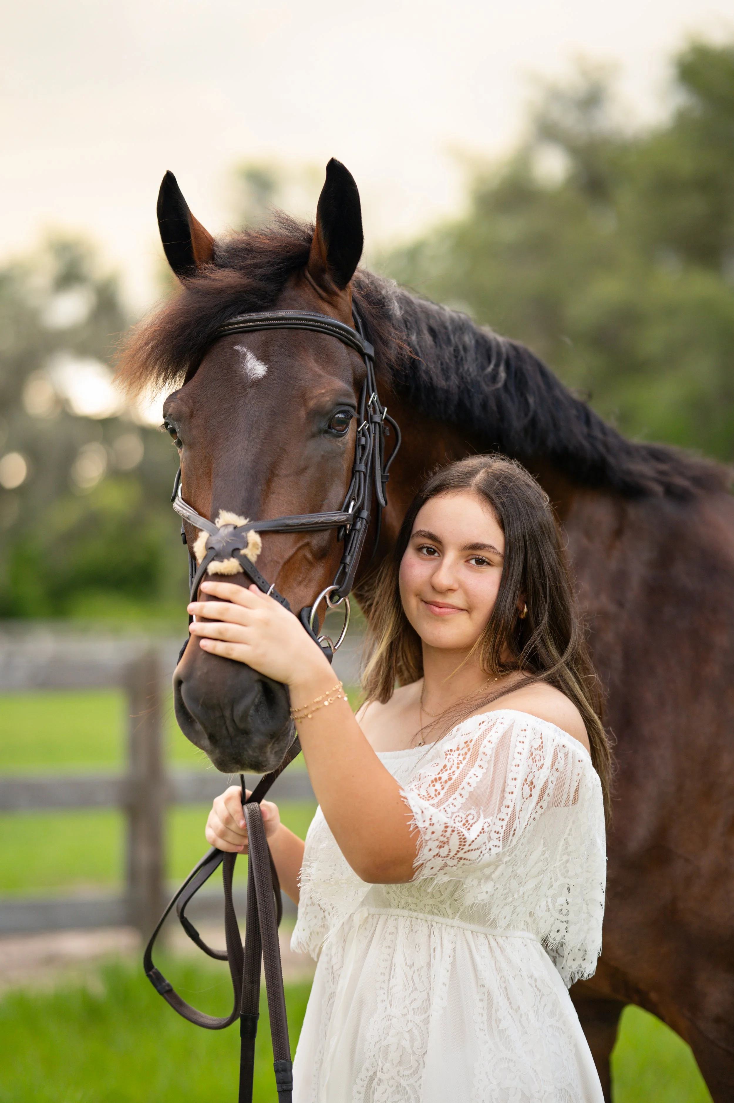 A young woman in a white lace dress holding a brown horse's bridle outdoors with trees and a wooden fence in the background.