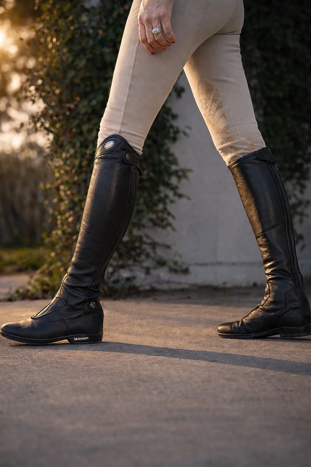 A person wearing beige pants and black knee-high riding boots standing outdoors on a paved surface, with greenery and a wall in the background.