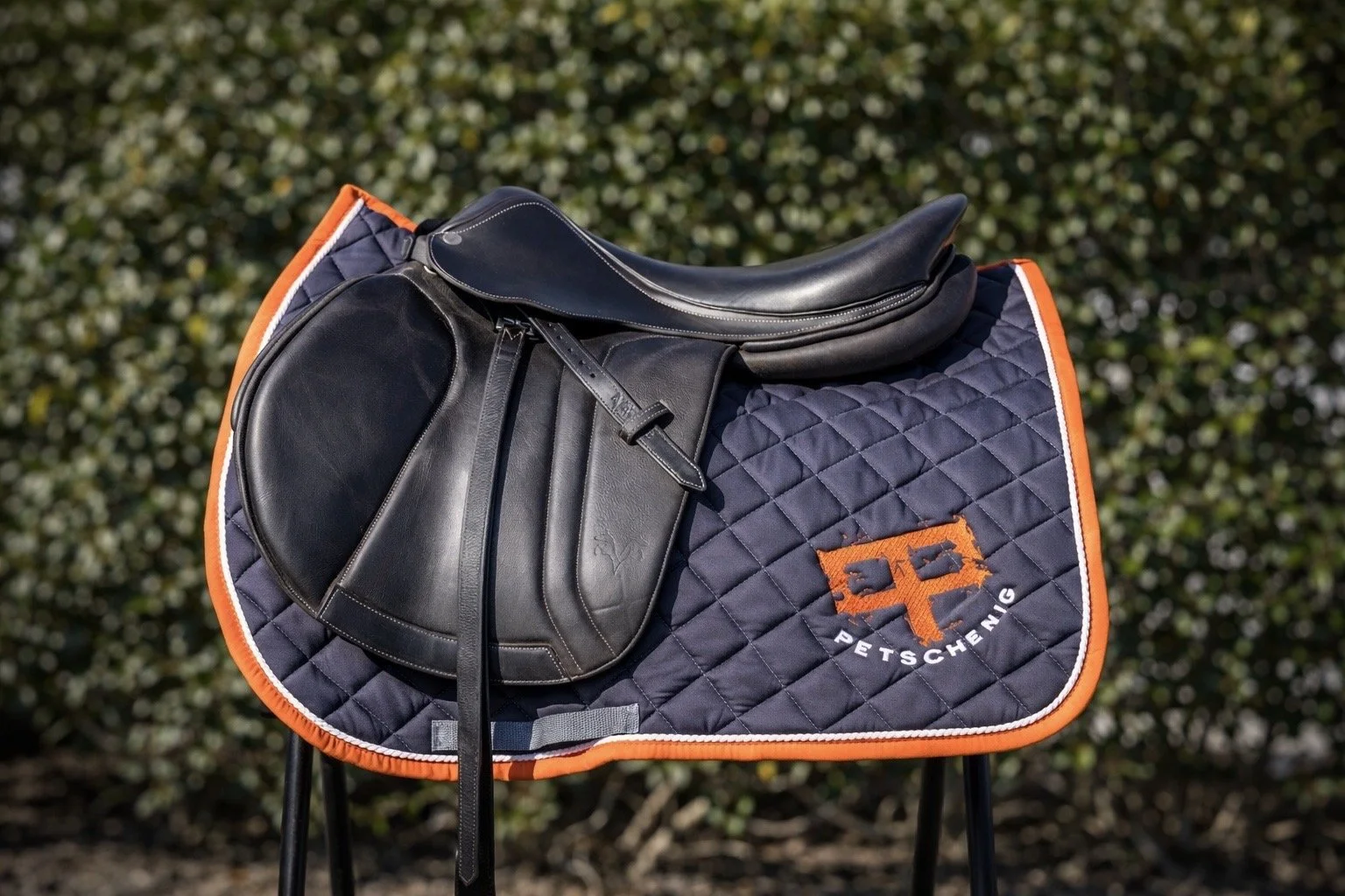 A black riding saddle on a padded saddle pad with an embroidered logo reading 'Petschenig' on a three-legged stand outdoors.