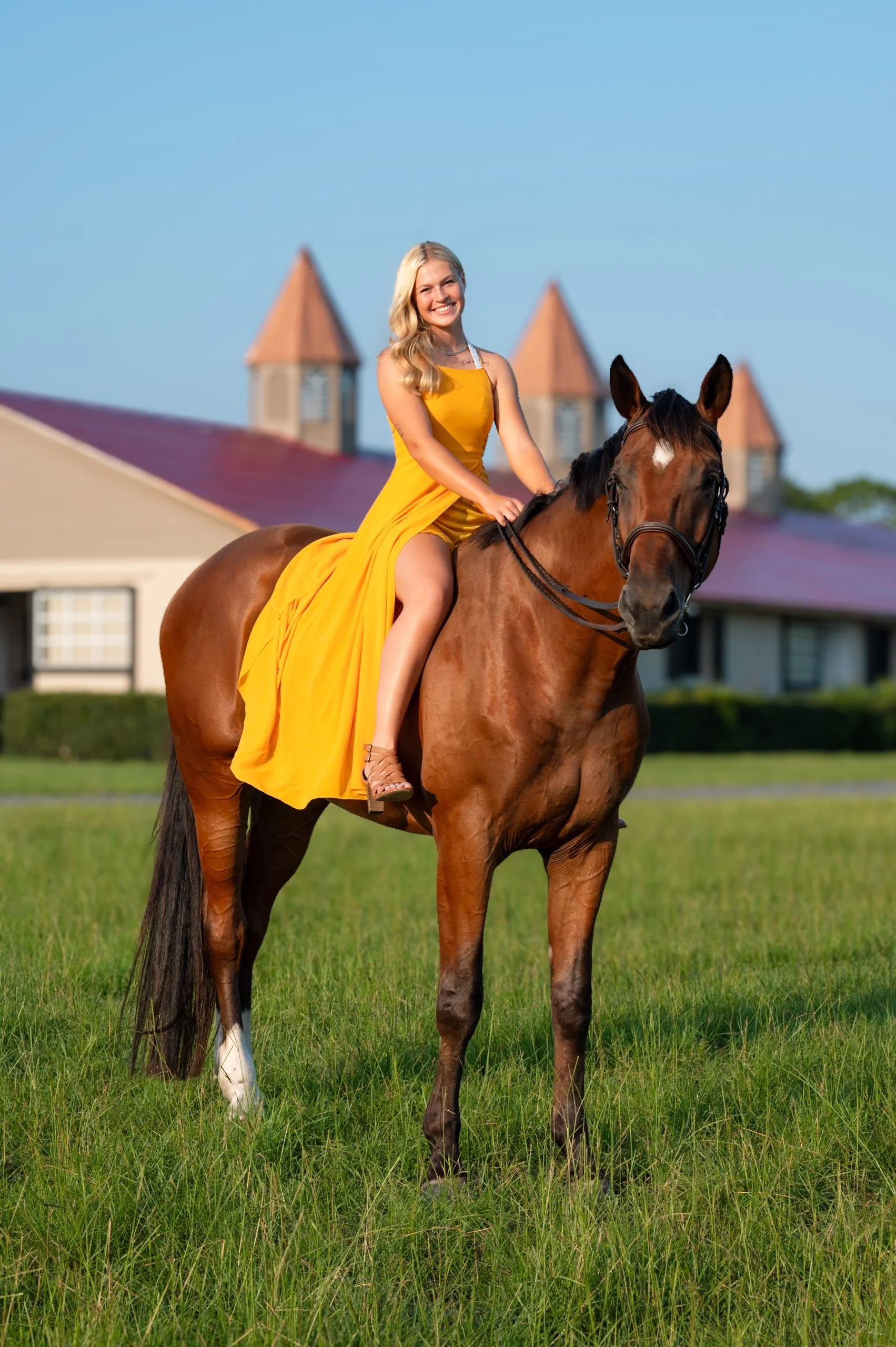 A young woman in a yellow dress riding a brown horse with a white marking on its face on a grassy field, with a building with red roofs in the background under a blue sky.