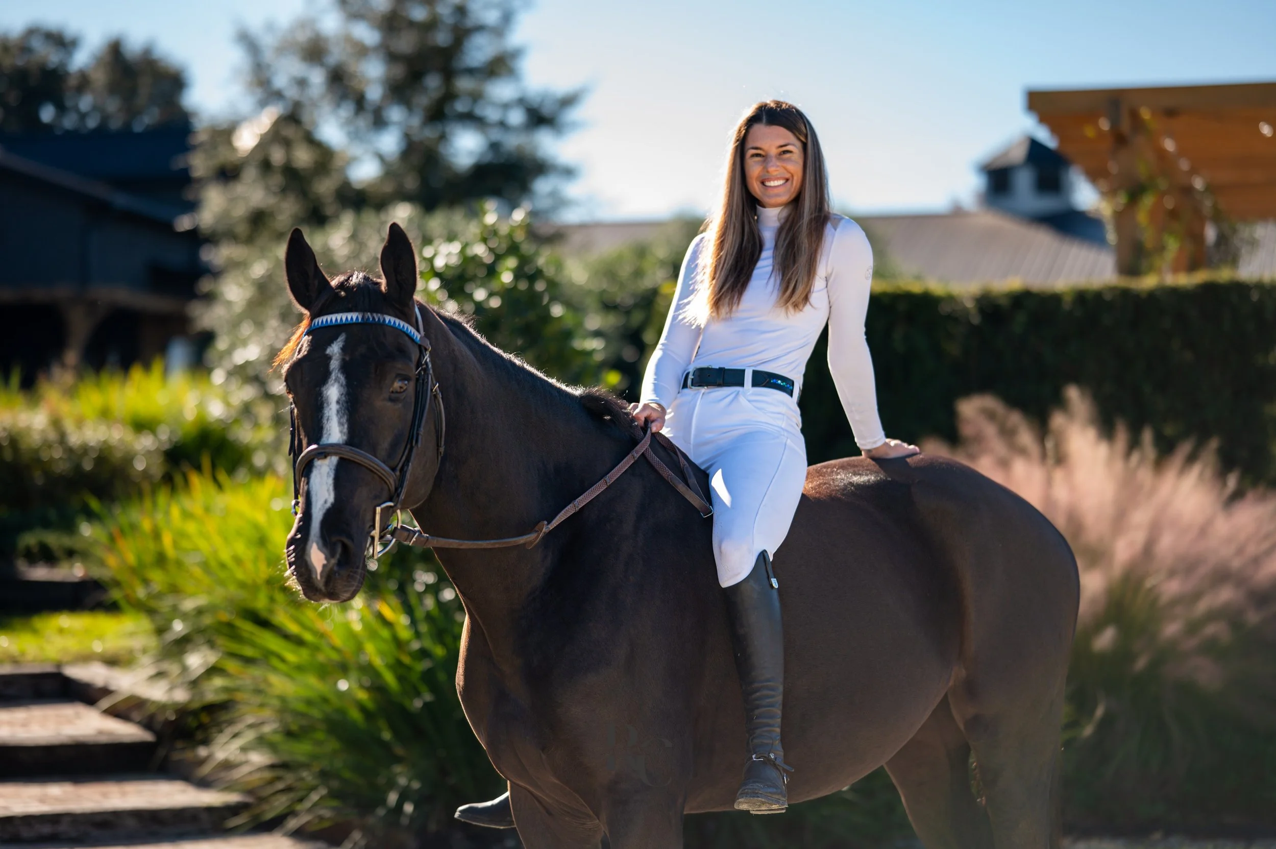 A woman riding a black horse outdoors on a sunny day, smiling and wearing a white outfit.