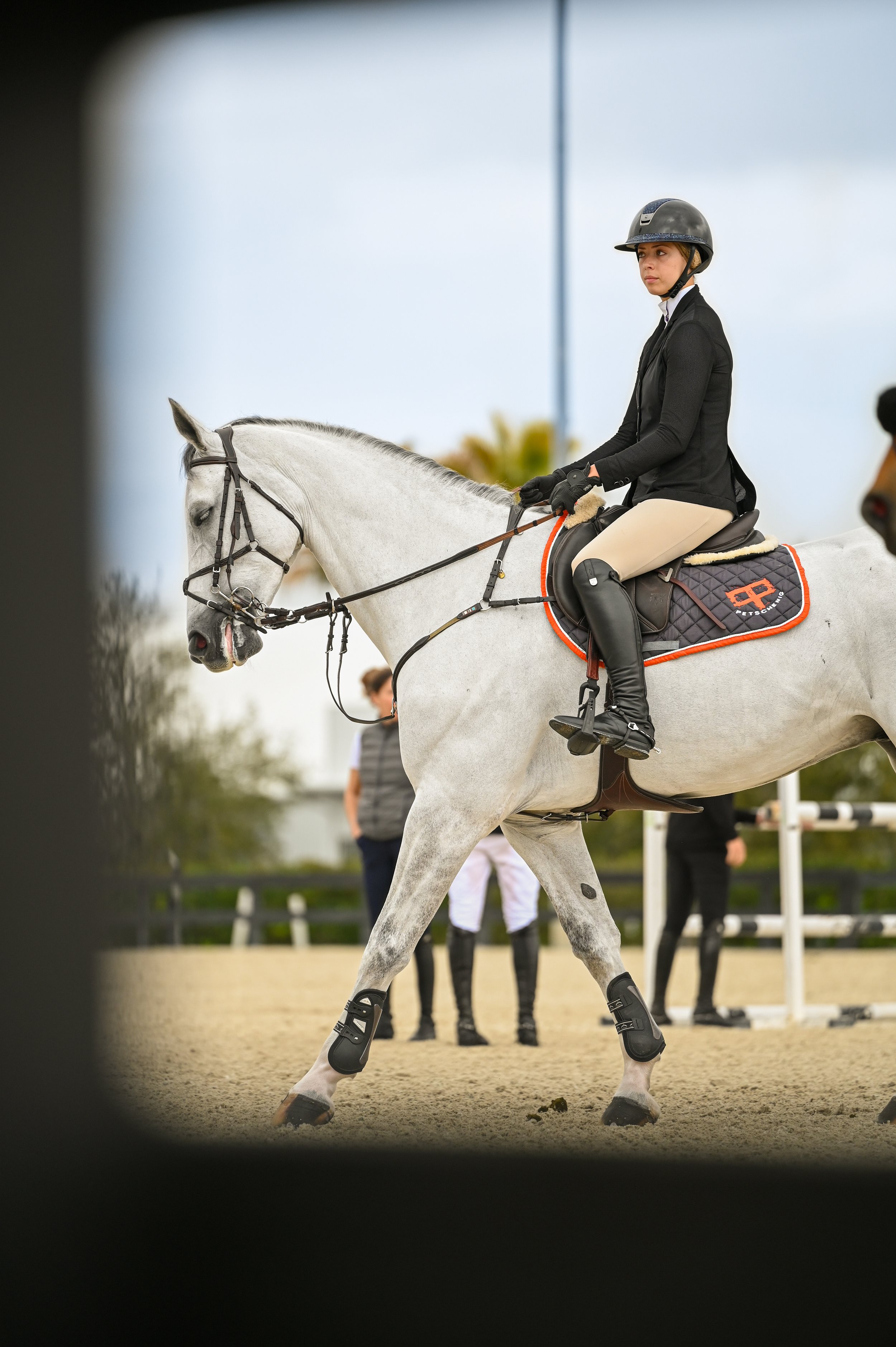 A woman riding a white horse during a horseback riding event, wearing a black helmet, black jacket, beige pants, and black riding boots, with other people in the background.