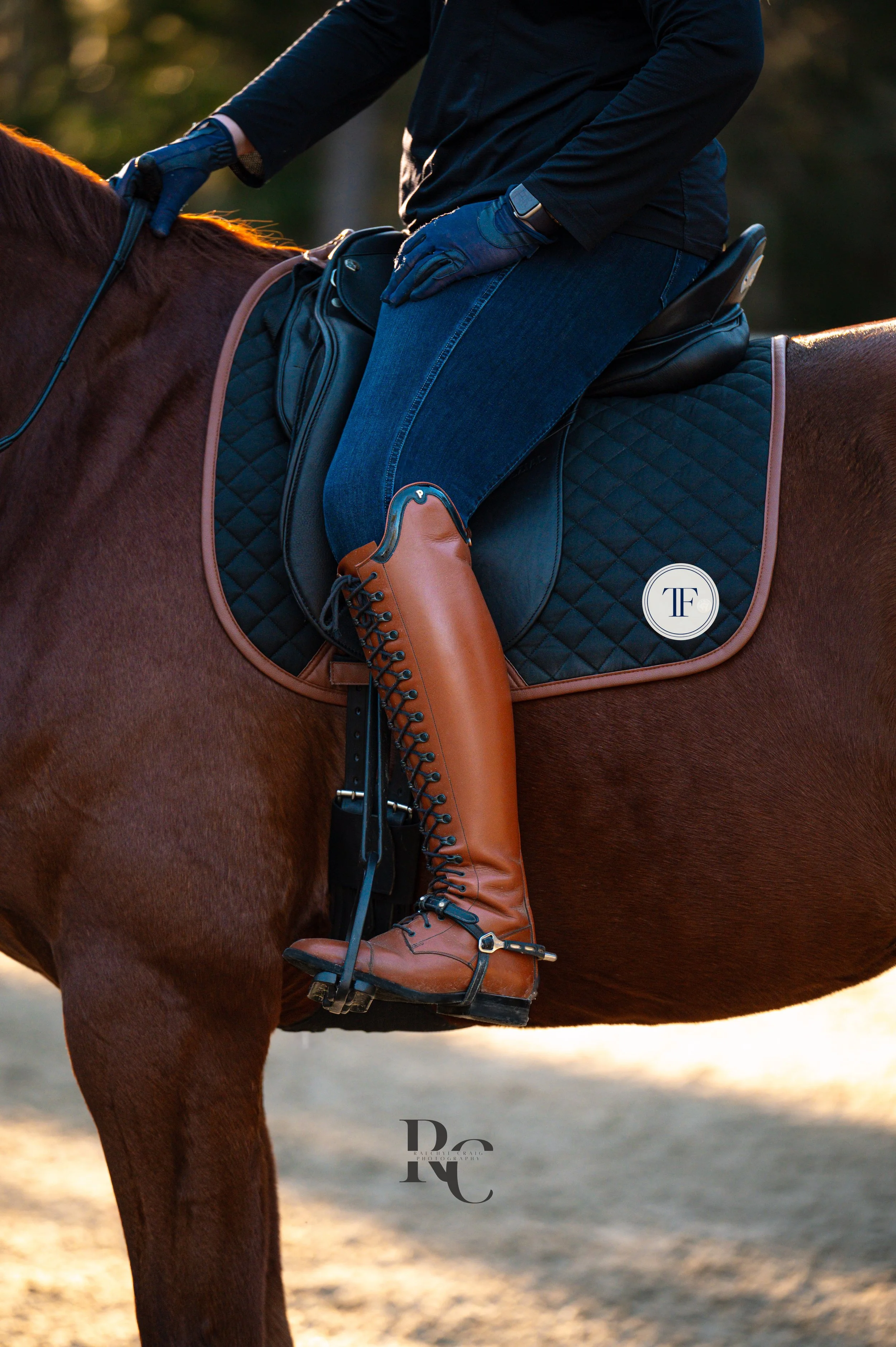 Close-up of a person riding a brown horse, wearing tan riding boots, dark blue jeans, a black jacket, and blue gloves, on a black quilted saddle pad with a white circular logo.