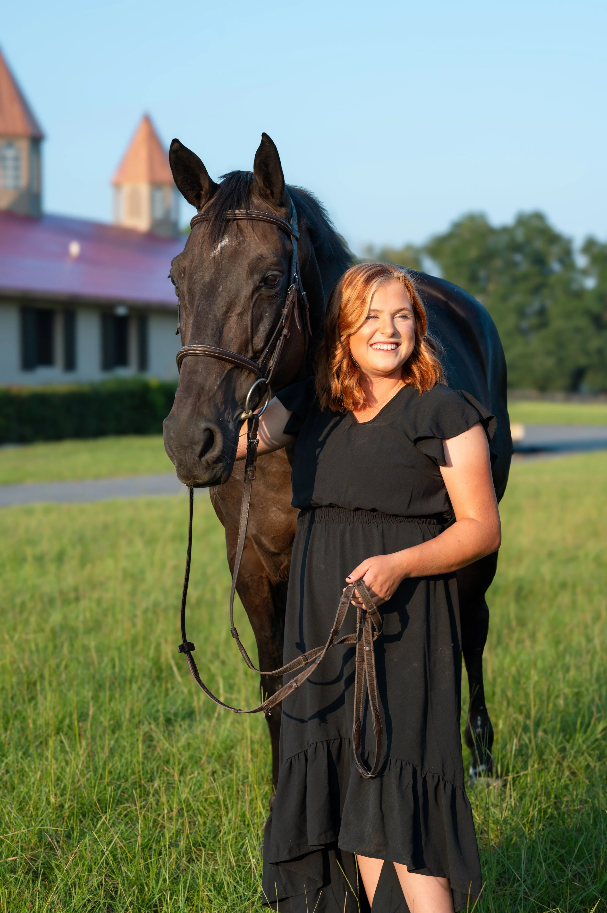 A woman in a black dress standing in a grassy field, smiling and holding the reins of a black horse, with a building and trees in the background on a sunny day.