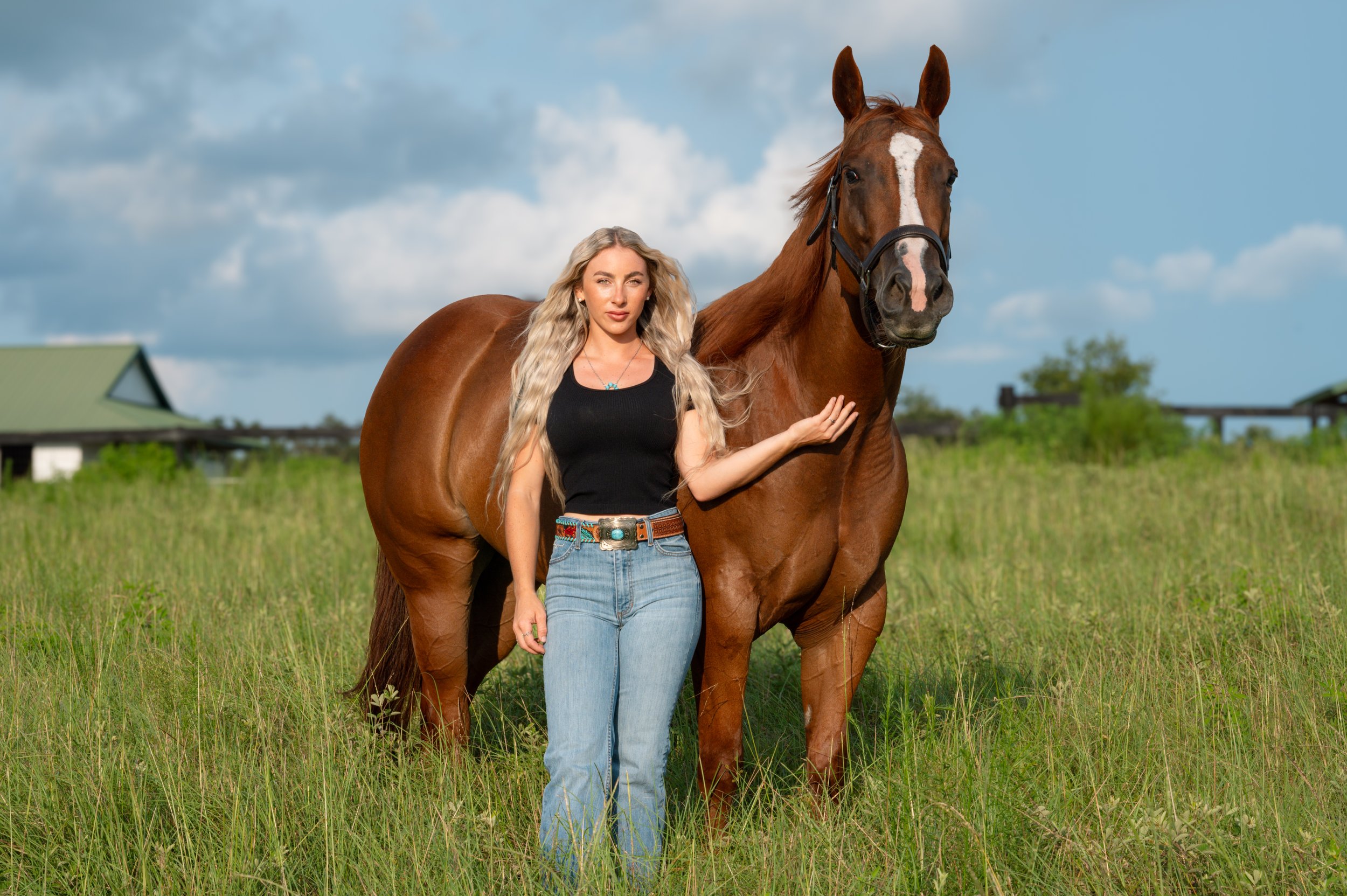 A woman with long blonde hair standing next to a brown horse with a white face marking in a grassy field under a partly cloudy sky.