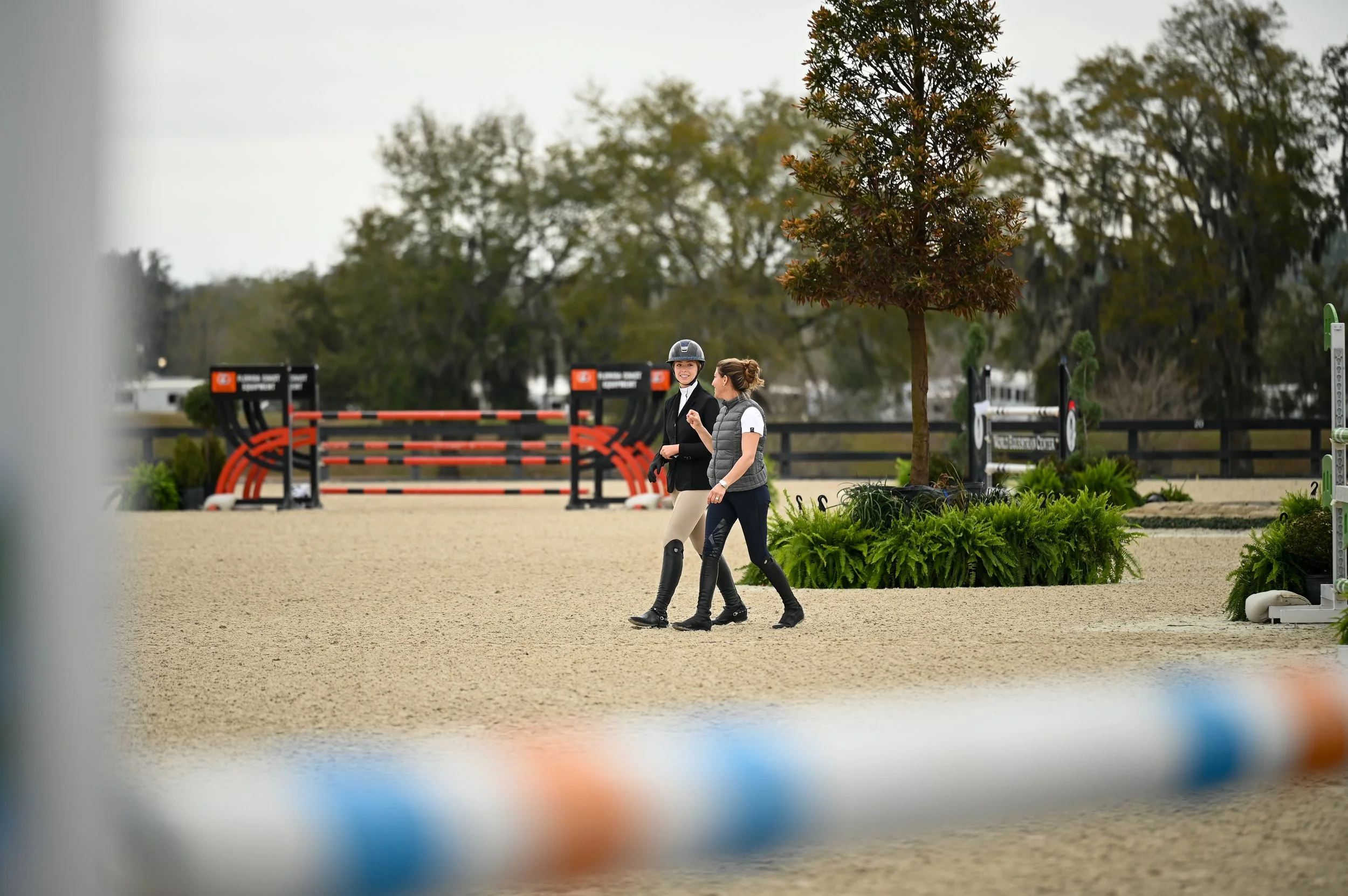 Two women walking and talking outdoors near a horse jumping arena, with a tree and greenery in the background.