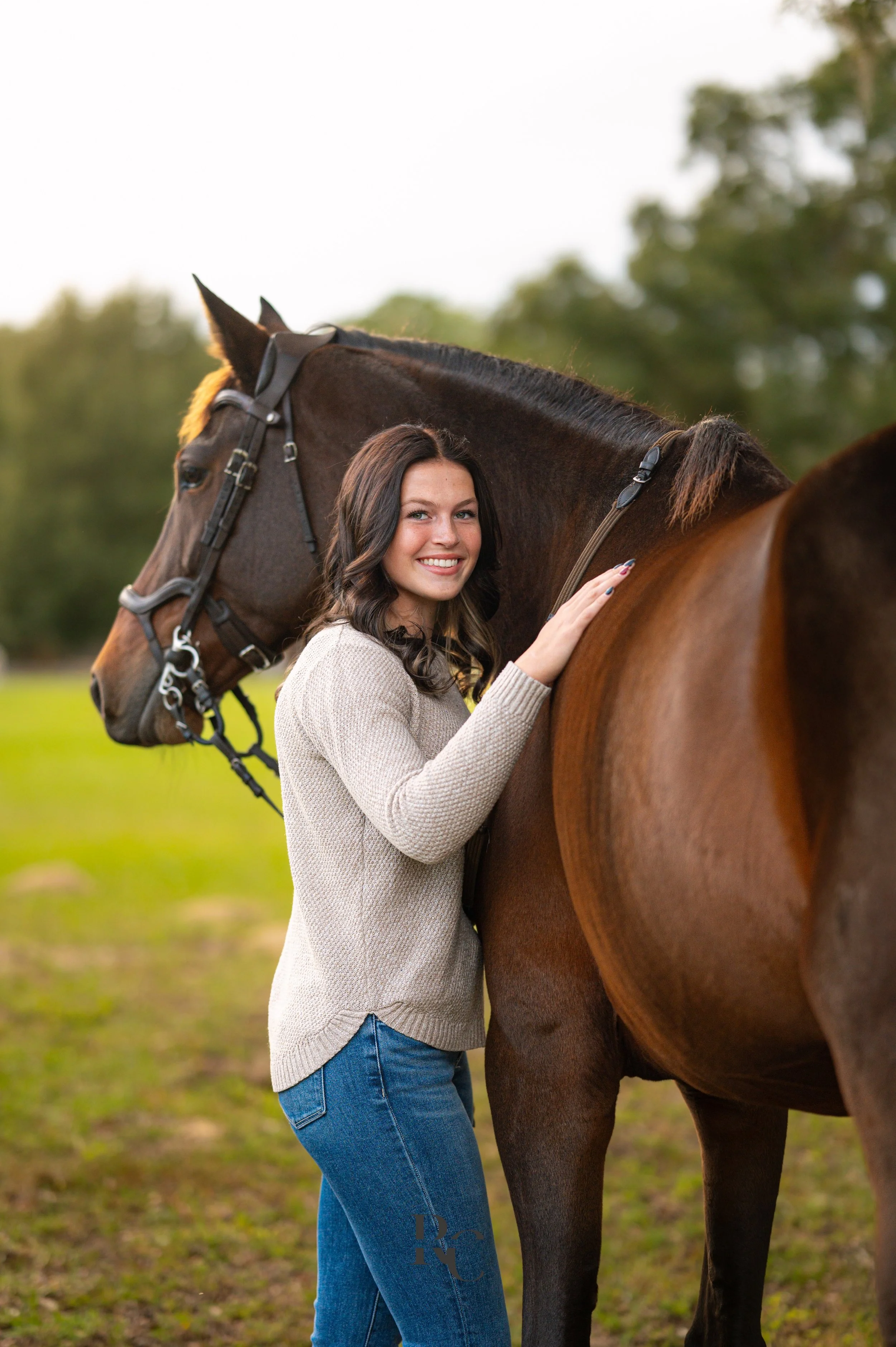 A young woman smiling and standing next to a brown horse, gently holding its neck with her left hand outdoors on a grassy field.