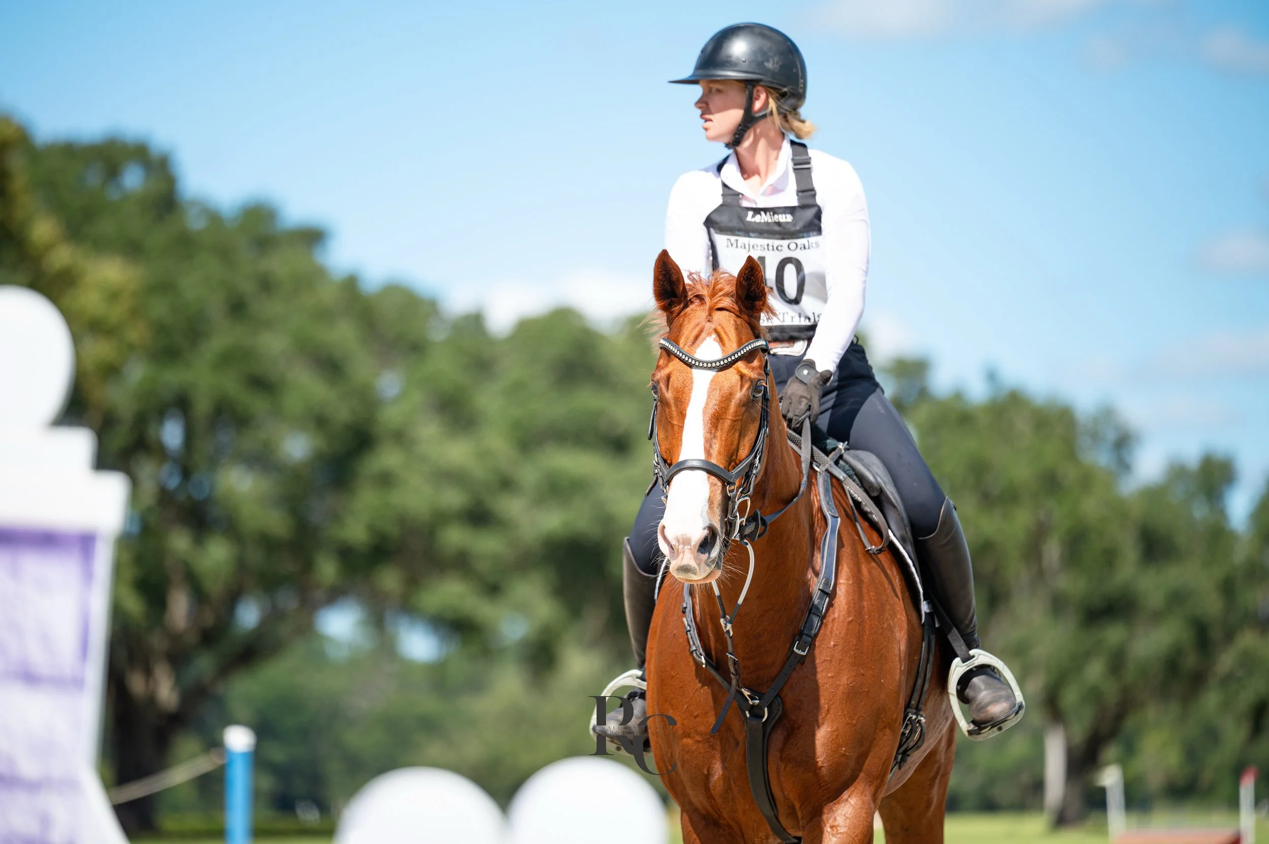 A woman riding a chestnut horse during a cross-country equestrian event, wearing a riding helmet, white shirt, and black riding gear with numbered bib.