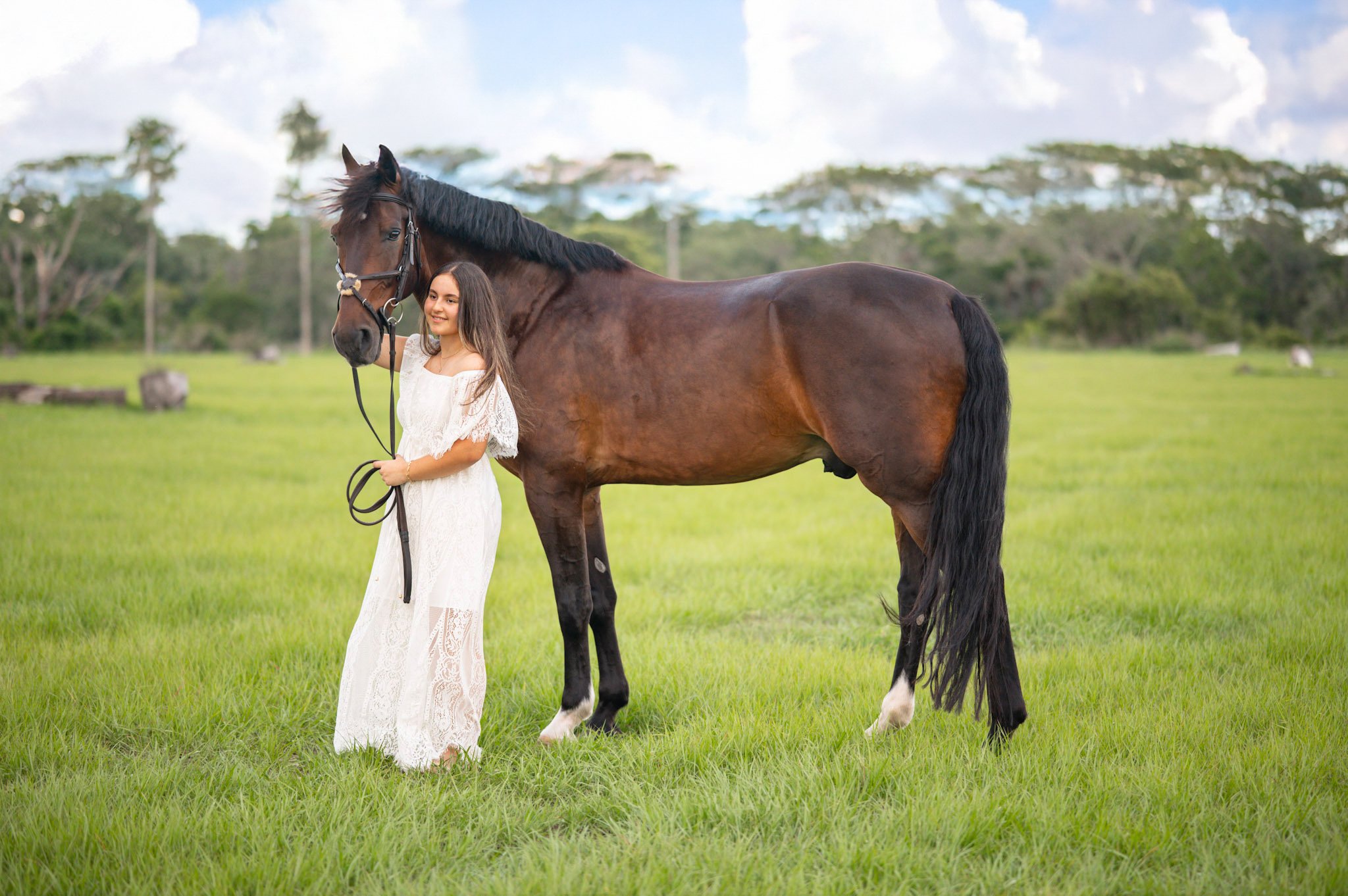 A girl in a white lace dress standing next to a brown horse in a grassy field with trees in the background.