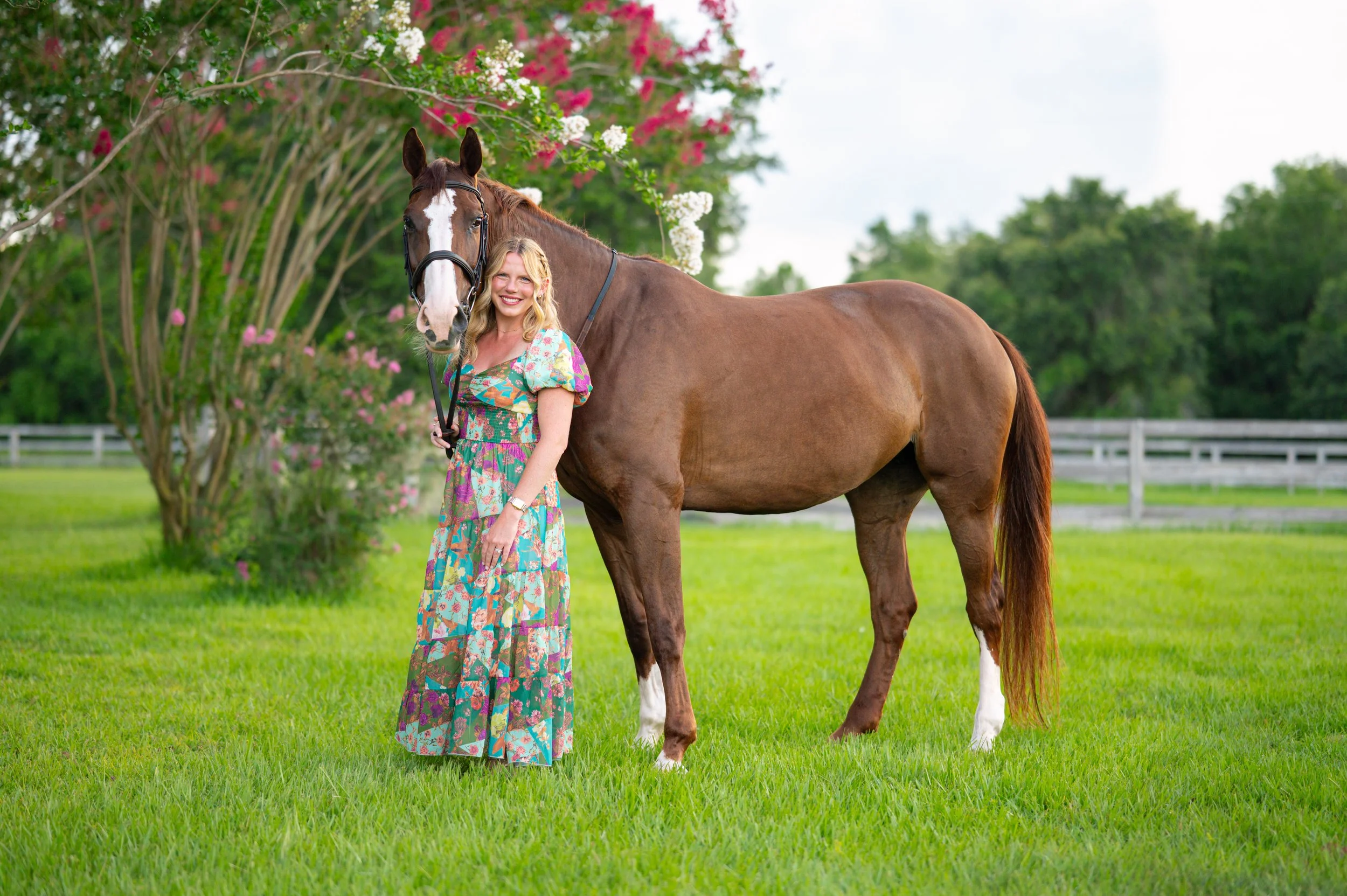 A young woman in a floral dress standing next to a brown and white horse on a green lawn with trees and a white fence in the background.