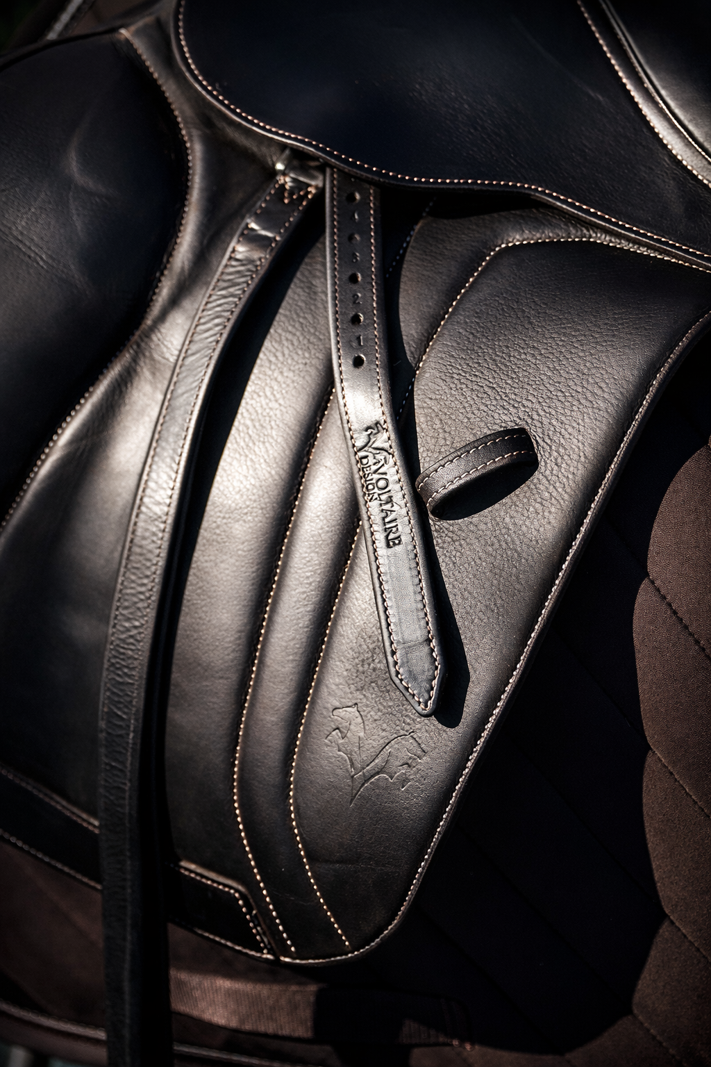 Close-up of a black golf bag with a gold-stitched logo and a leather strap, resting on a brown cushioned surface.