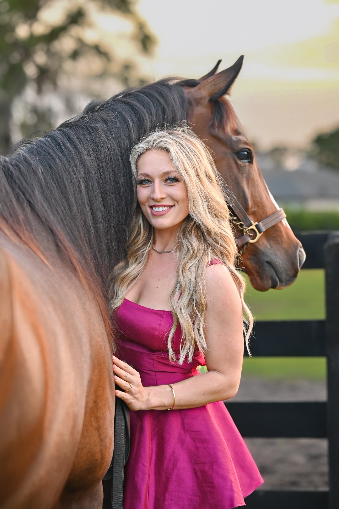 A young woman with long blonde hair in a pink dress stands next to a brown horse with a black mane, smiling at the camera outdoors.