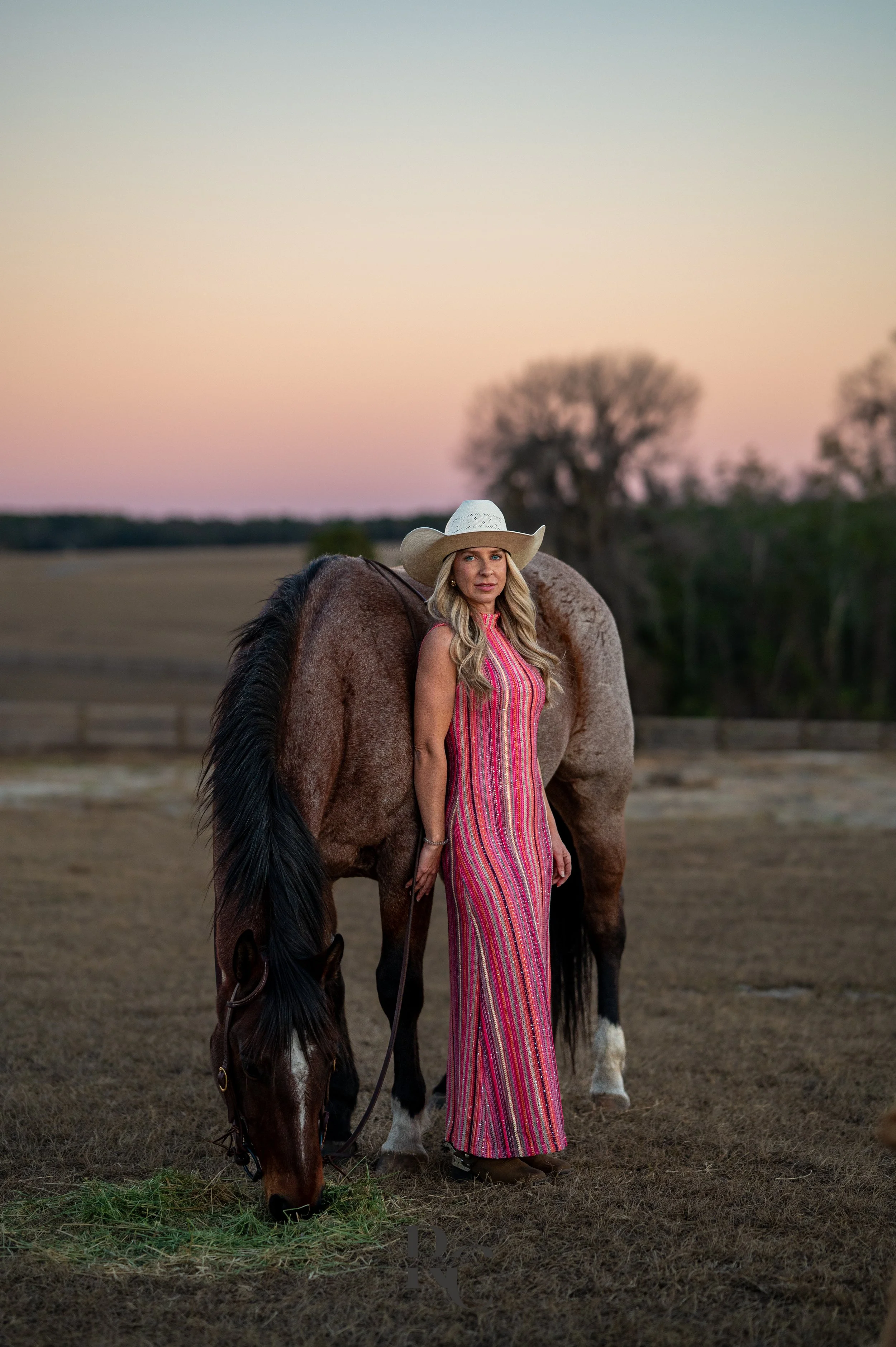 A woman in a colorful striped dress and a wide-brimmed cowboy hat stands next to a brown horse with a black mane in an open field during sunset, with trees in the background.