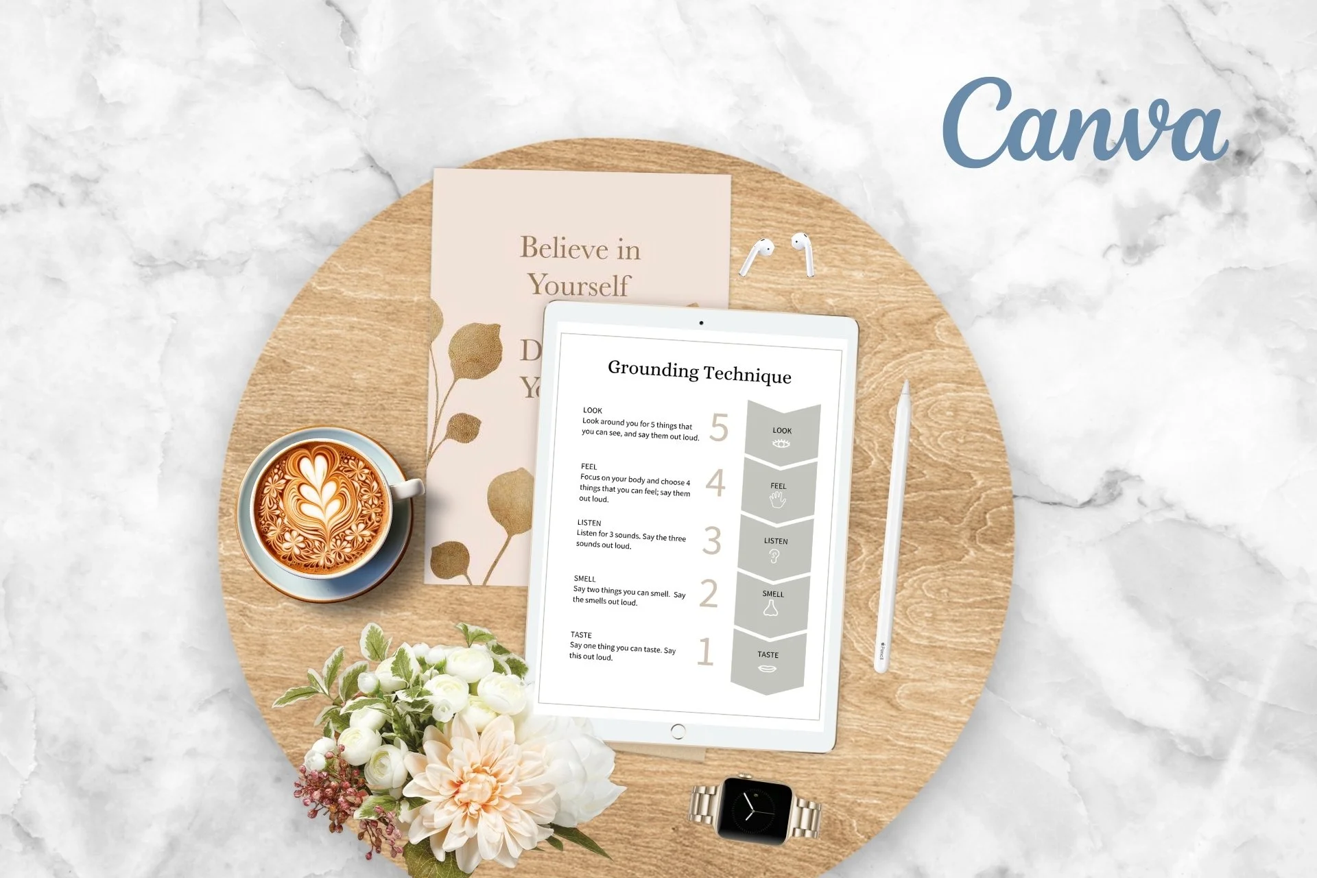 A round wooden table on a marble surface holding a tablet displaying a grounding technique chart, a coffee cup with latte art, a small bouquet of white and pink flowers, a white pen, a silver wristwatch, and a piece of paper with motivational text.