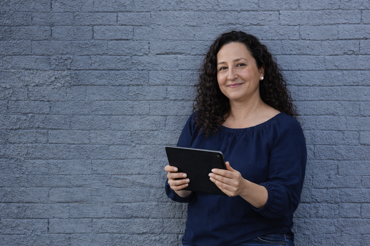 A woman with dark curly hair, wearing a blue top and pearl earrings, is smiling while holding a black tablet in front of a textured gray brick wall.