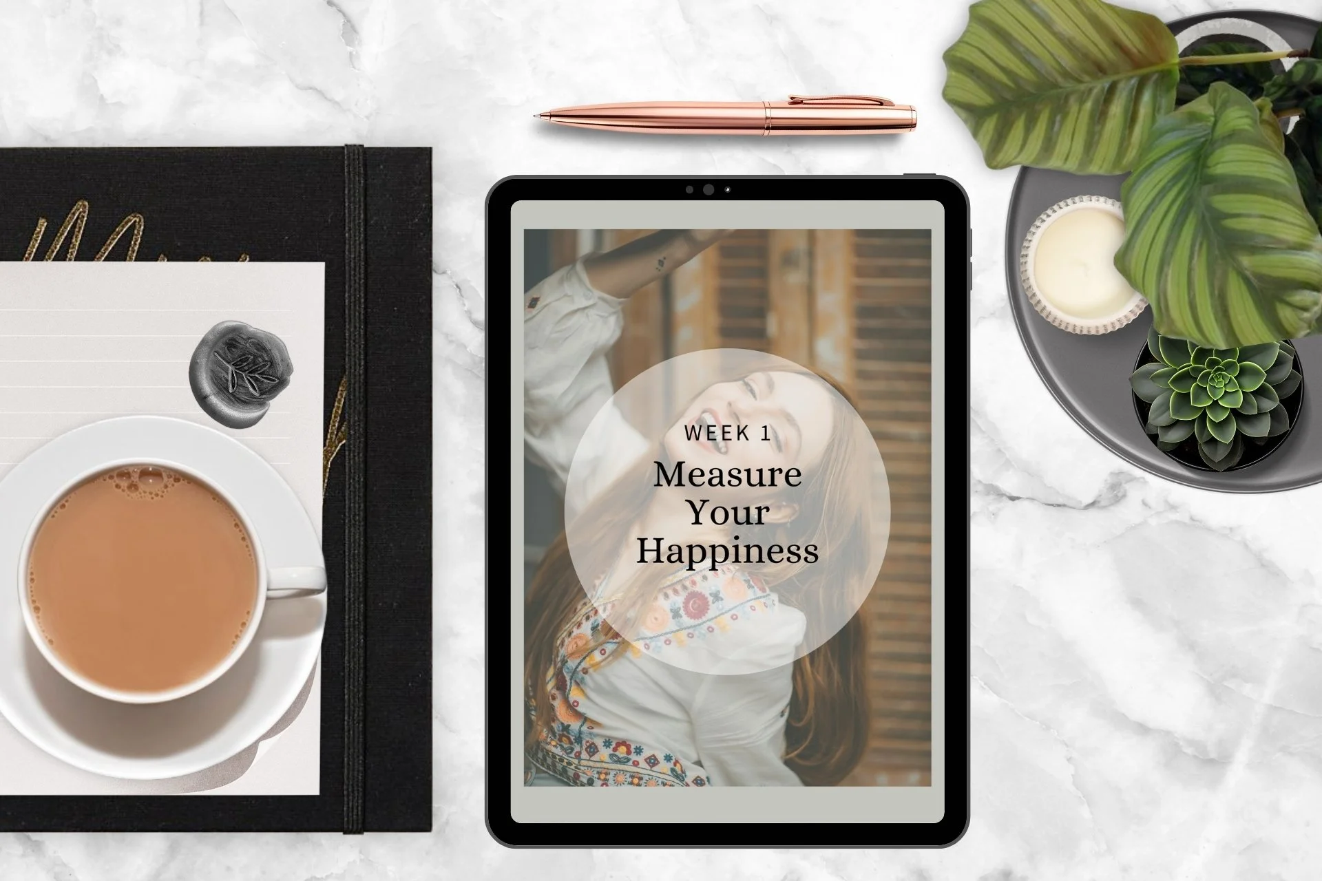 A flat lay of a workspace with a tablet displaying 'Week 1 Measure Your Happiness,' a cup of tea, a notebook with a paper and pen, and a potted plant on a marble surface.