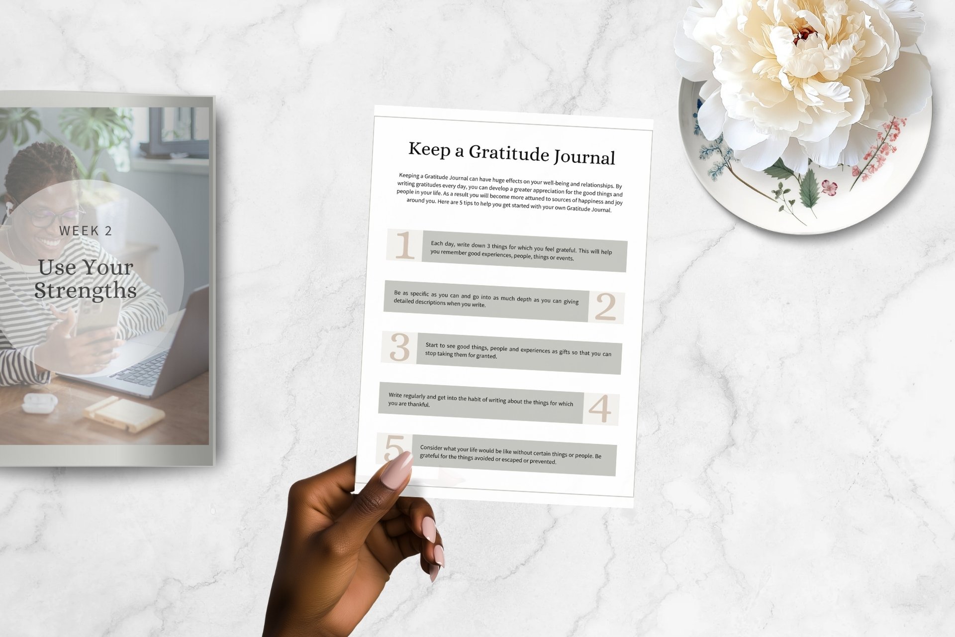 A person's hand holding a printed gratitude journal page on a white marble surface, with a photo of a woman smiling and using her phone, a white flower in a vase, and a tablet or laptop on the desk.