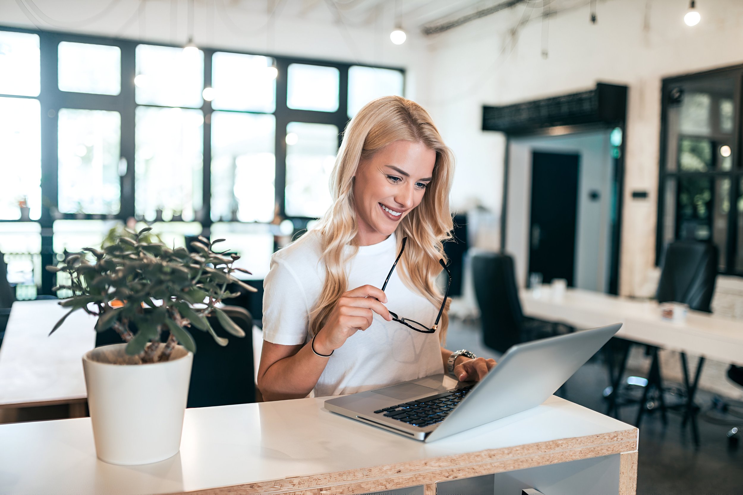 A woman with blonde hair smiling and looking at her laptop while holding her glasses in her hand in a modern office or cafe setting with large windows, plants, and tables.