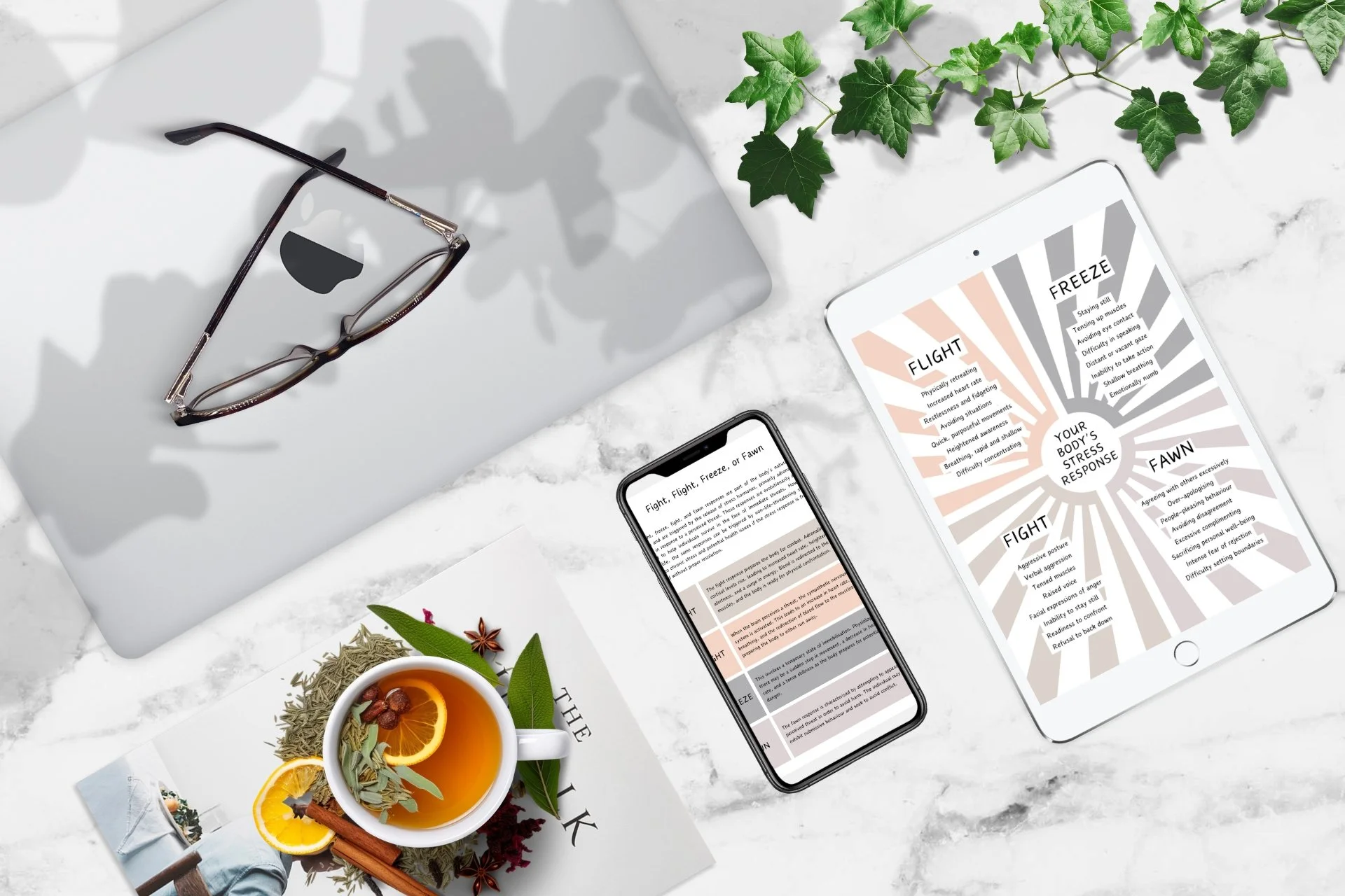 A flat lay of a workspace on a white marble surface, including a closed silver laptop with eyeglasses on top, an iPad displaying stress response information with a radial diagram, an iPhone with matching text and infographic, a teacup of herbal tea with lemon slices and spices on a magazine, and a green ivy plant in the corner.