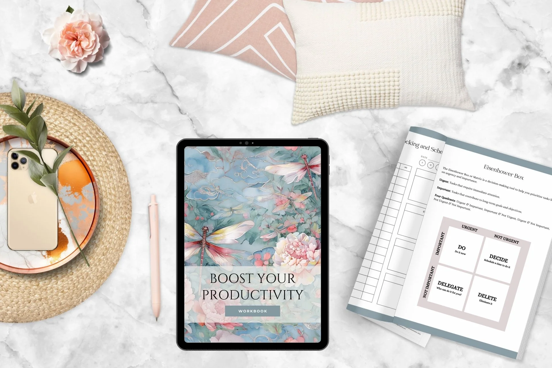 The image shows a flat lay of productivity and workspace items on a marble surface, including a tablet with a floral background and the text 'Boost Your Productivity Workbook,' a smartphone on a woven coaster with a branch and green leaves, a pink pen, a light-colored pillow, a pink and white pillow, a stack of papers with schedule and task lists titled 'Eisenhower Box,' a pink flower, and a floral-patterned cloth.