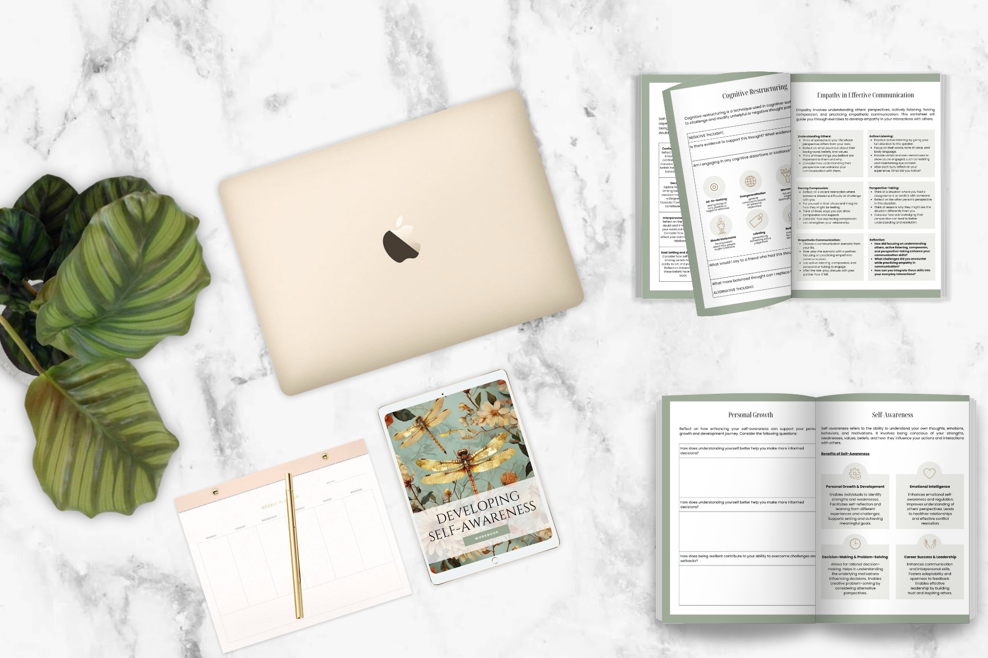 A silver MacBook laptop, a white and gold weekly planner, a framed digital book titled 'Developing Self-Awareness,' and an open booklet on personal growth are placed on a white marble surface, with a bunch of large green leaves on the left side.