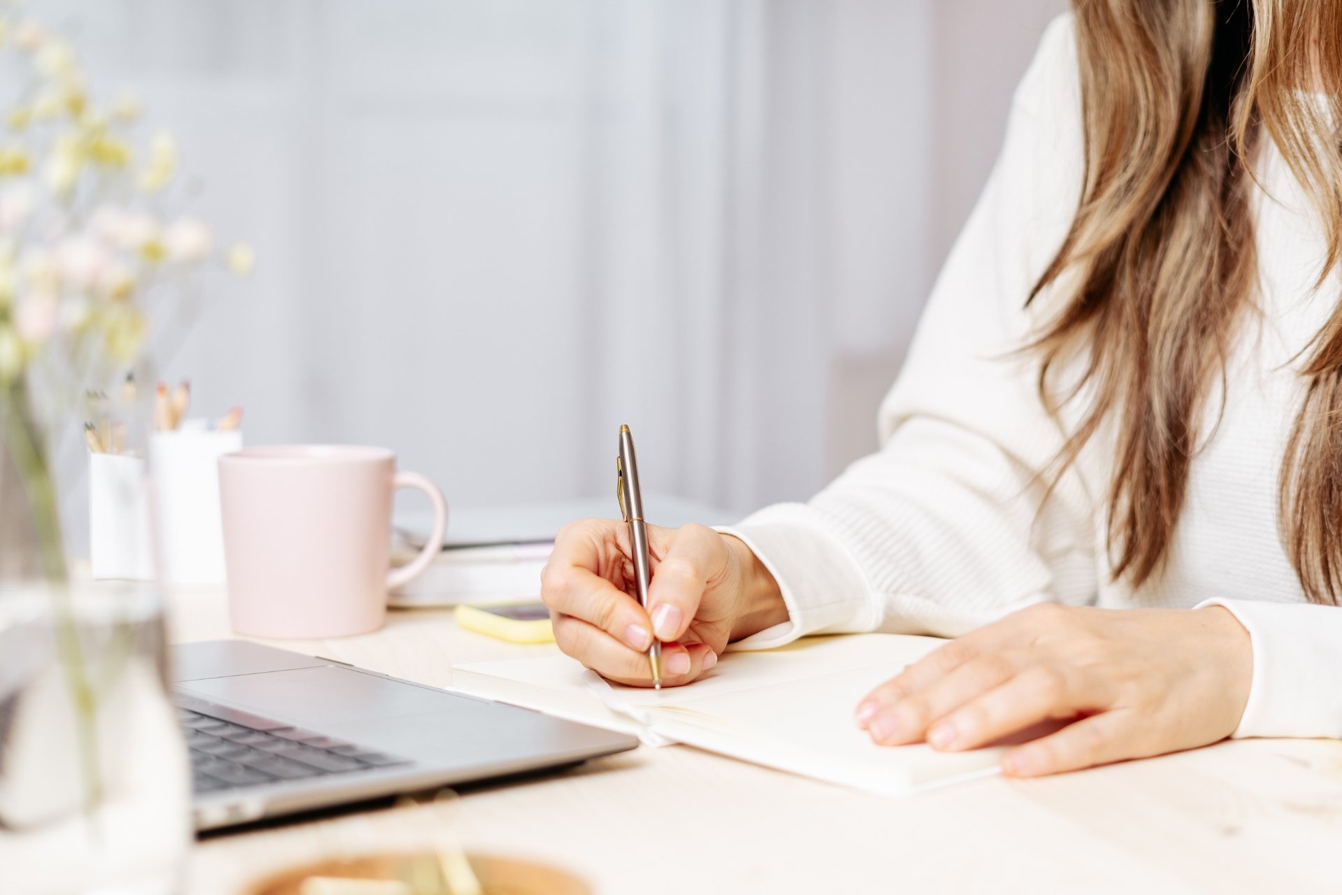 A woman writing in a notebook at a desk with a laptop, pink mug, and flowers.