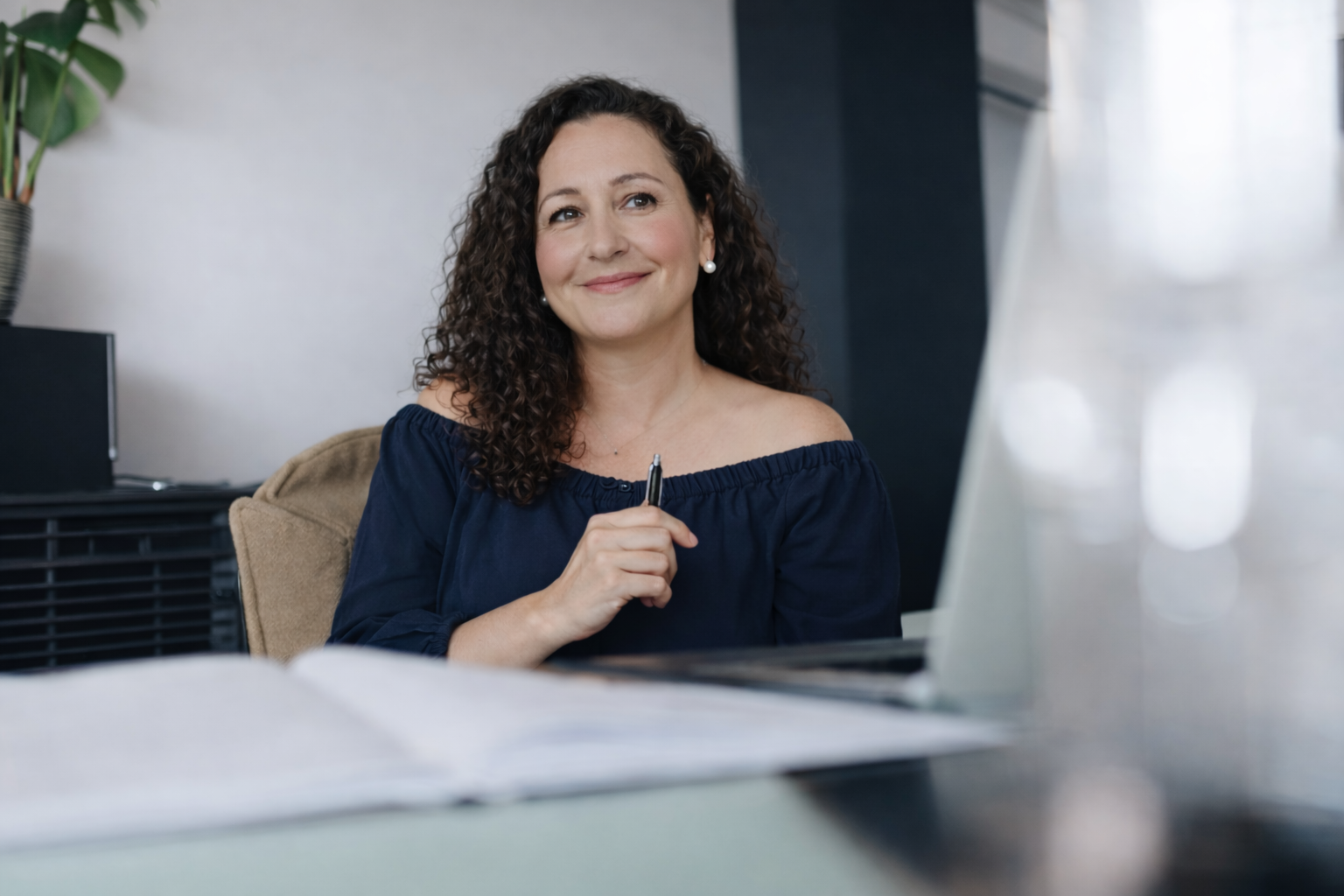 A woman with curly brown hair, wearing a navy blue off-shoulder top and pearl earrings, sitting at a desk in an office, smiling and holding a pen.