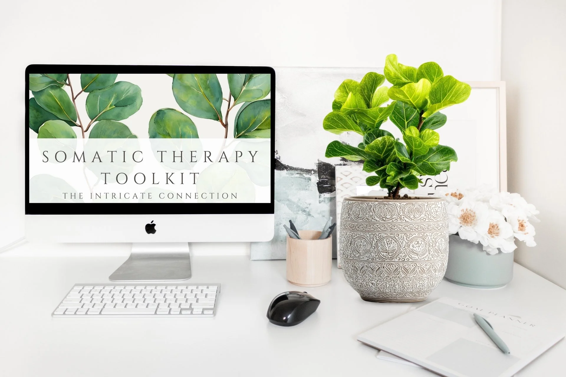 A computer screen displays a webpage titled 'Somatic Therapy Toolkit: The Intricate Connection' with a leafy plant background. The desk has a wireless keyboard, a mouse, a notepad with a pen, and plants with white flowers.