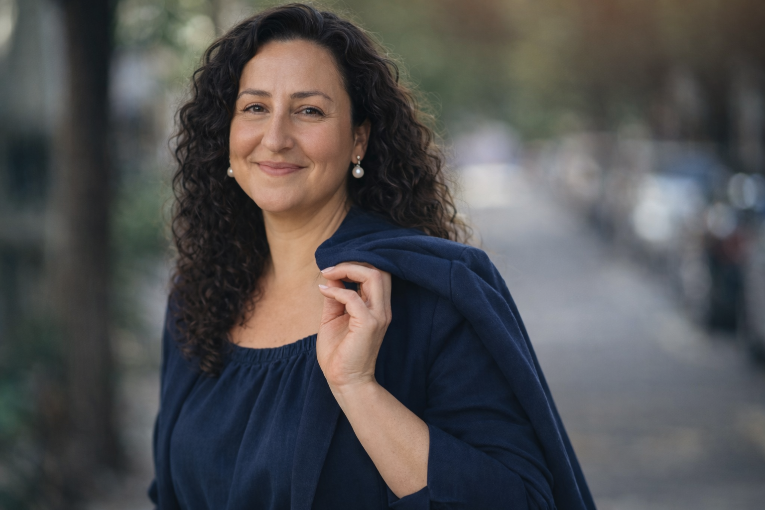 Smiling woman with curly dark hair wearing pearl earrings and a navy blue ensemble, holding her jacket over her shoulder on a tree-lined street.