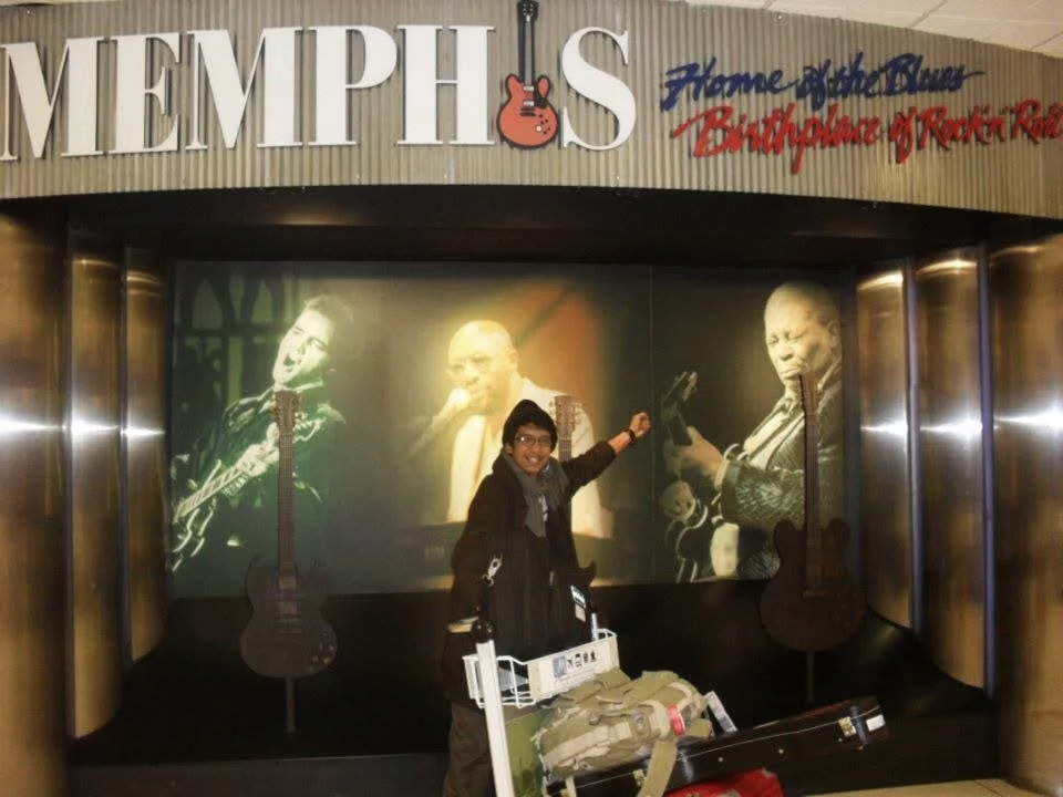 Person smiling and pointing at a Memphis display of a black-and-white photo of blues musicians with guitars, at an airport or similar location.