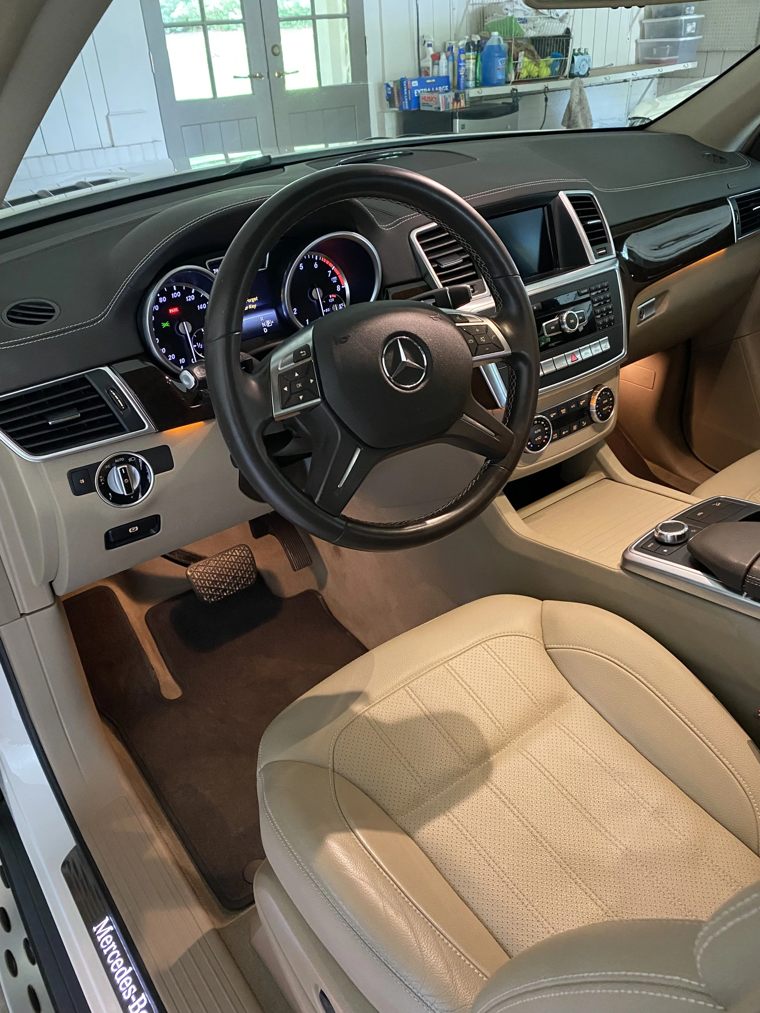 Interior of a Mercedes-Benz SUV featuring a leather steering wheel, dashboard with digital displays, beige leather seats, and center console controls.