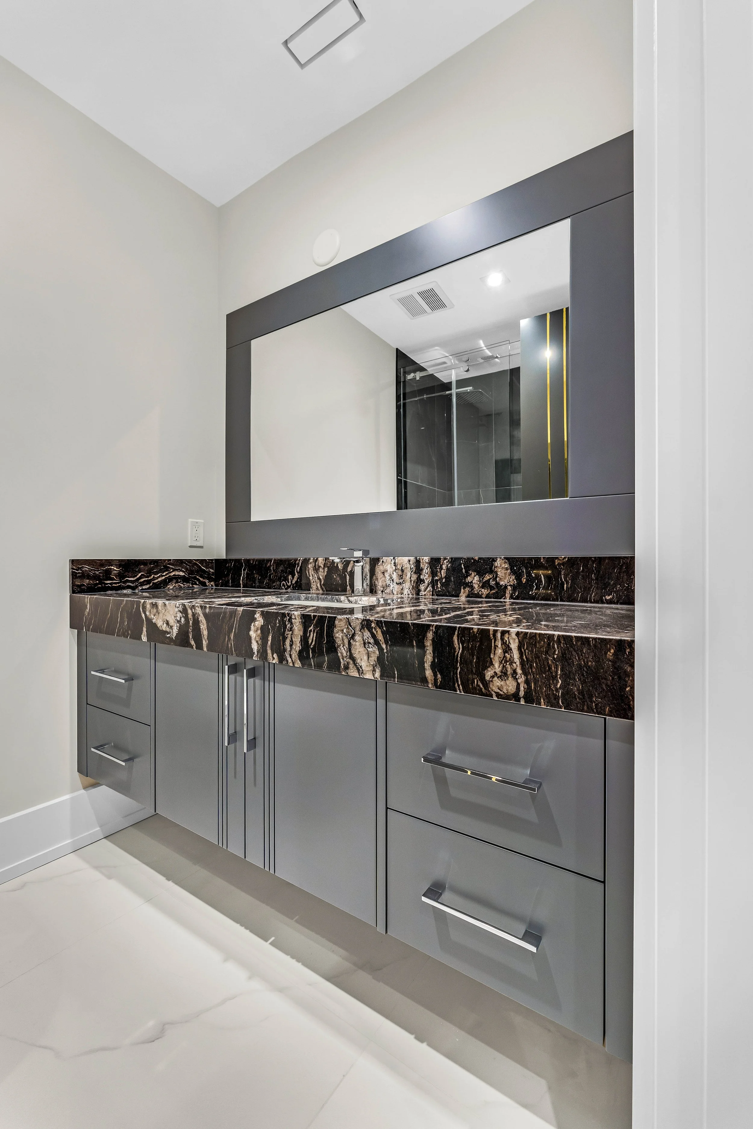 A modern bathroom vanity with a dark brown marble countertop and a grey cabinet below. A large rectangular mirror with a black frame is mounted above the sink. The wall behind the vanity is plain and off-white, and the floor is light-colored tile.