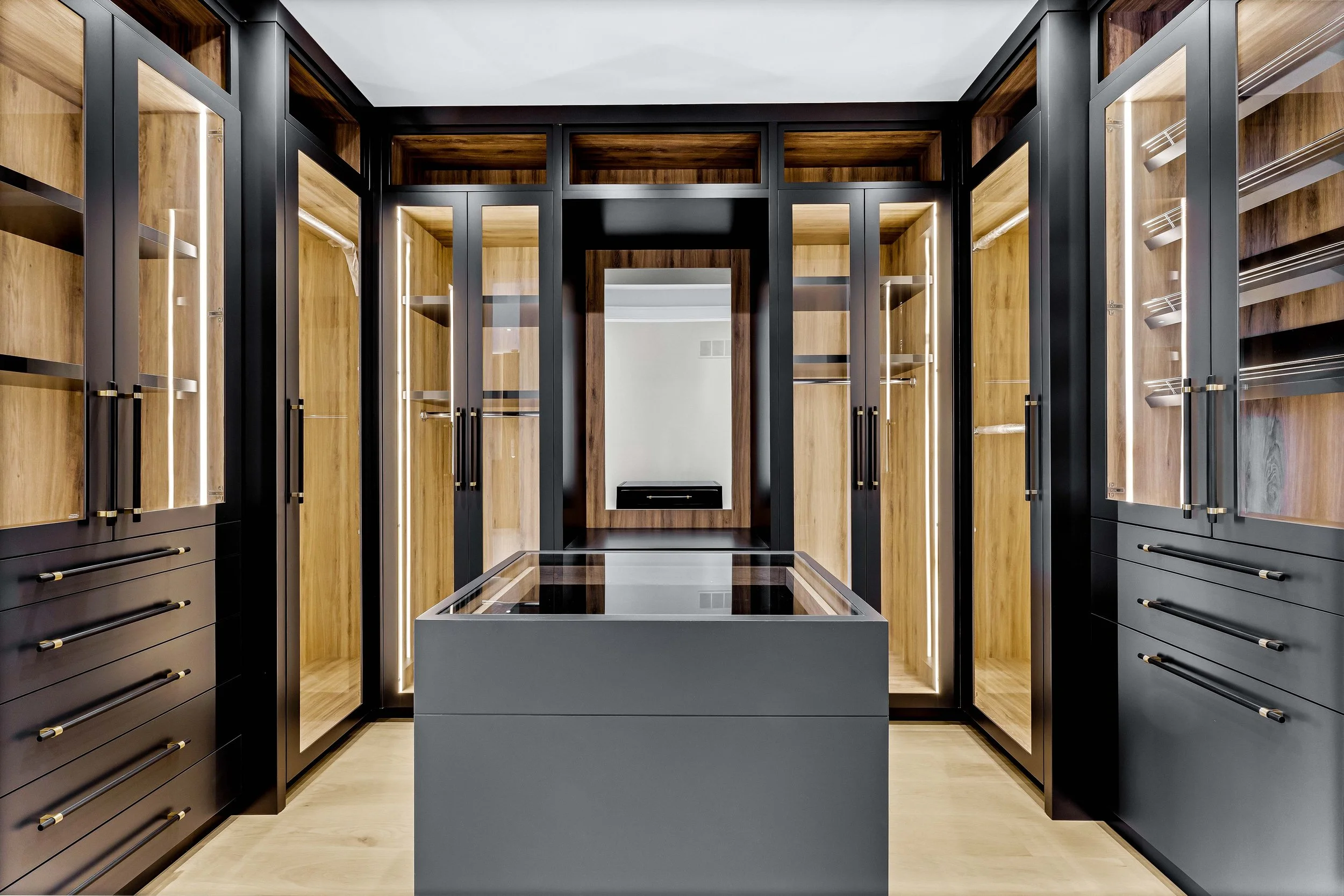 A modern walk-in closet with black and wood cabinetry, glass display cases, and a center glass-top island.