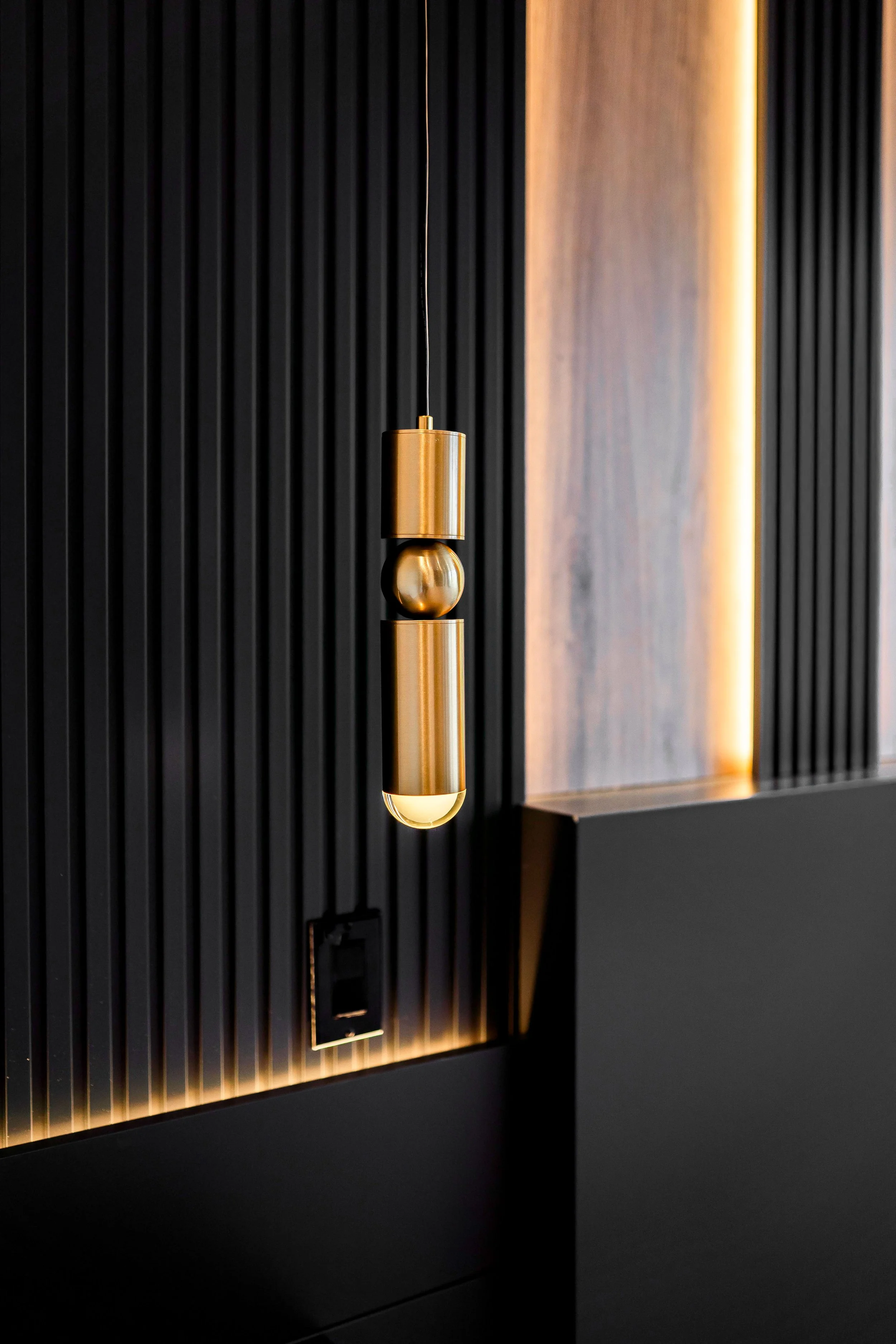 Modern pendant light fixture with a gold finish hanging in front of a textured black and wood wall with LED strip lighting.