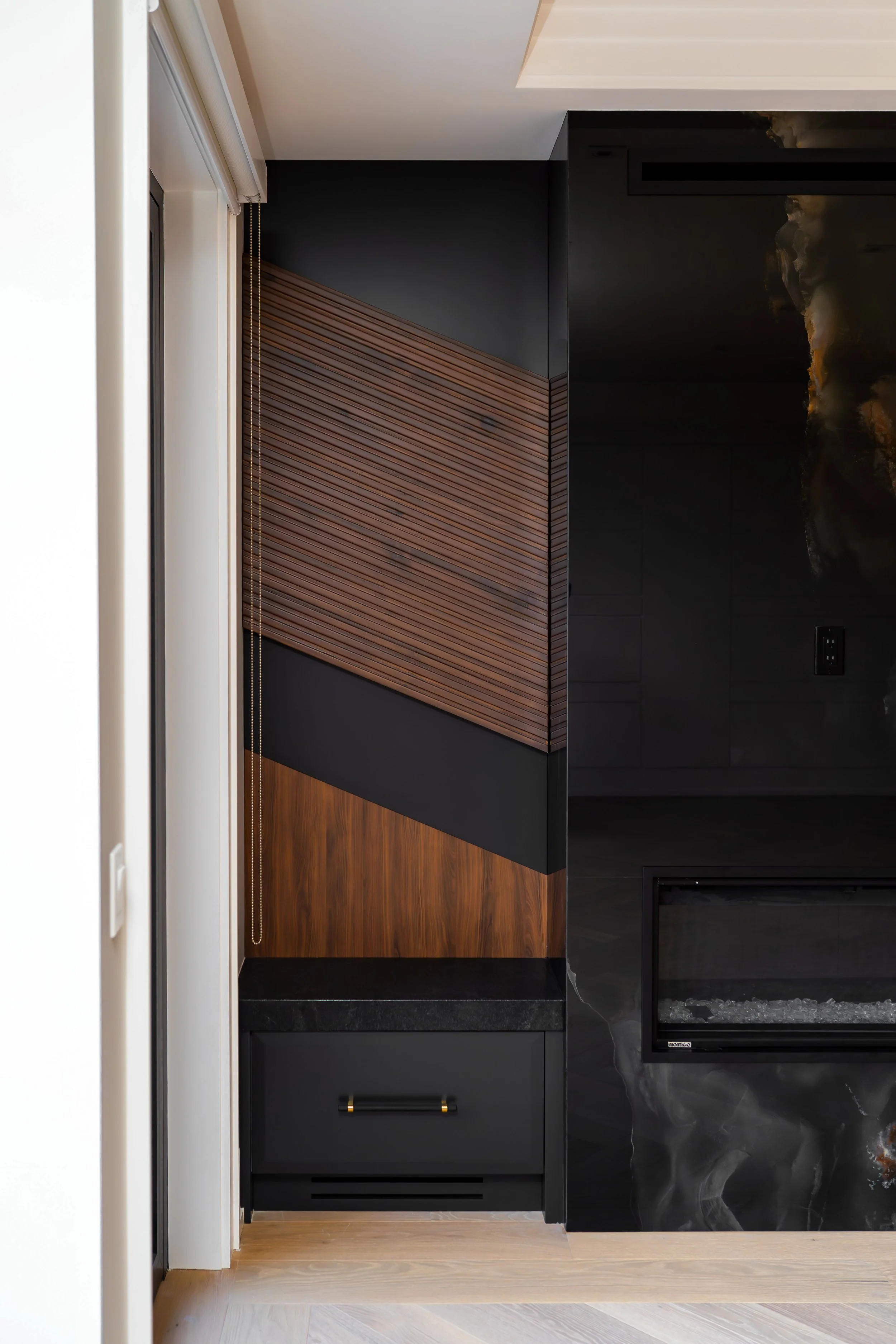 Interior view of a modern living space featuring a black and wooden accent wall next to a fireplace with a marble-like finish, and a small black cabinet with a handle at the bottom.