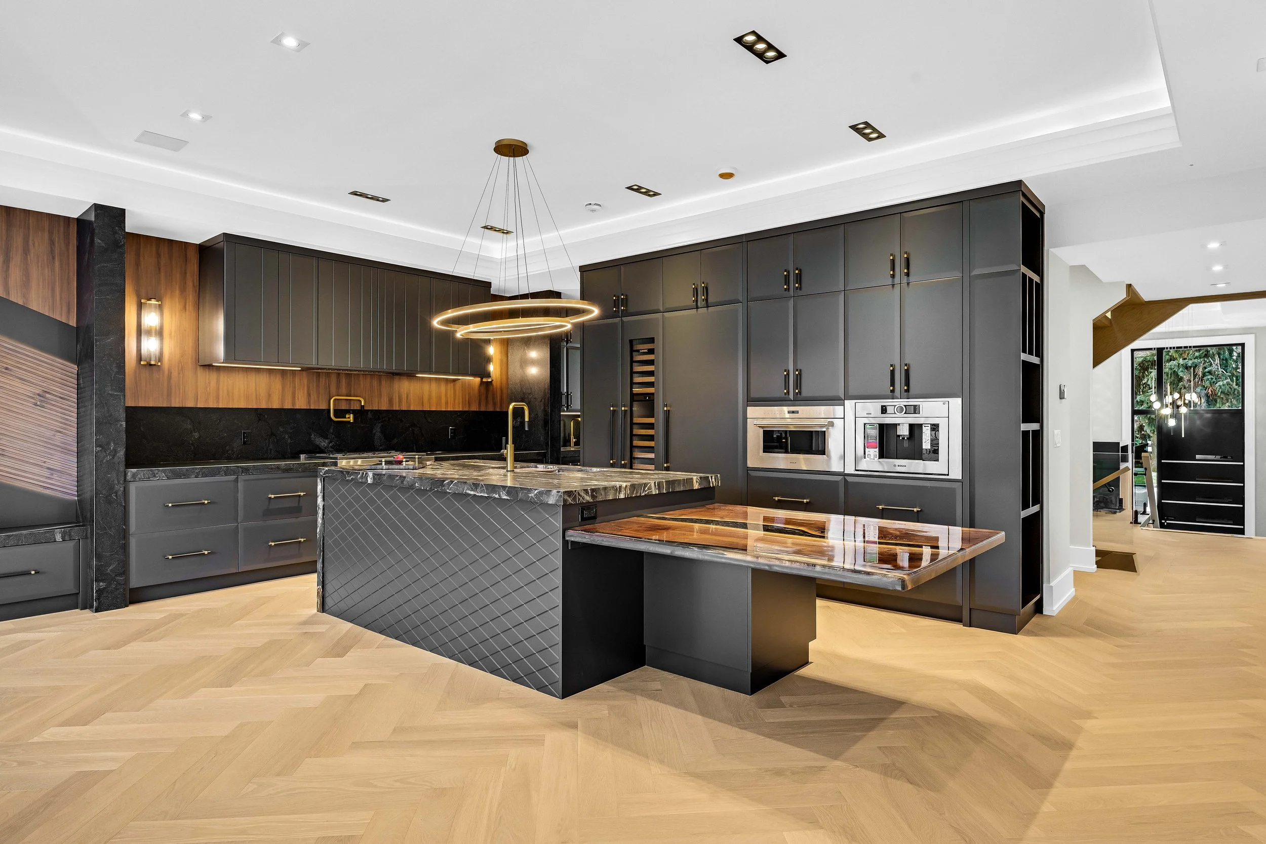 Modern kitchen with dark gray cabinets, black marble countertops, wooden accents, a large island with a patterned black base and marble top, and built-in stainless steel appliances. Light wood herringbone floors and contemporary lighting fixtures are also visible.
