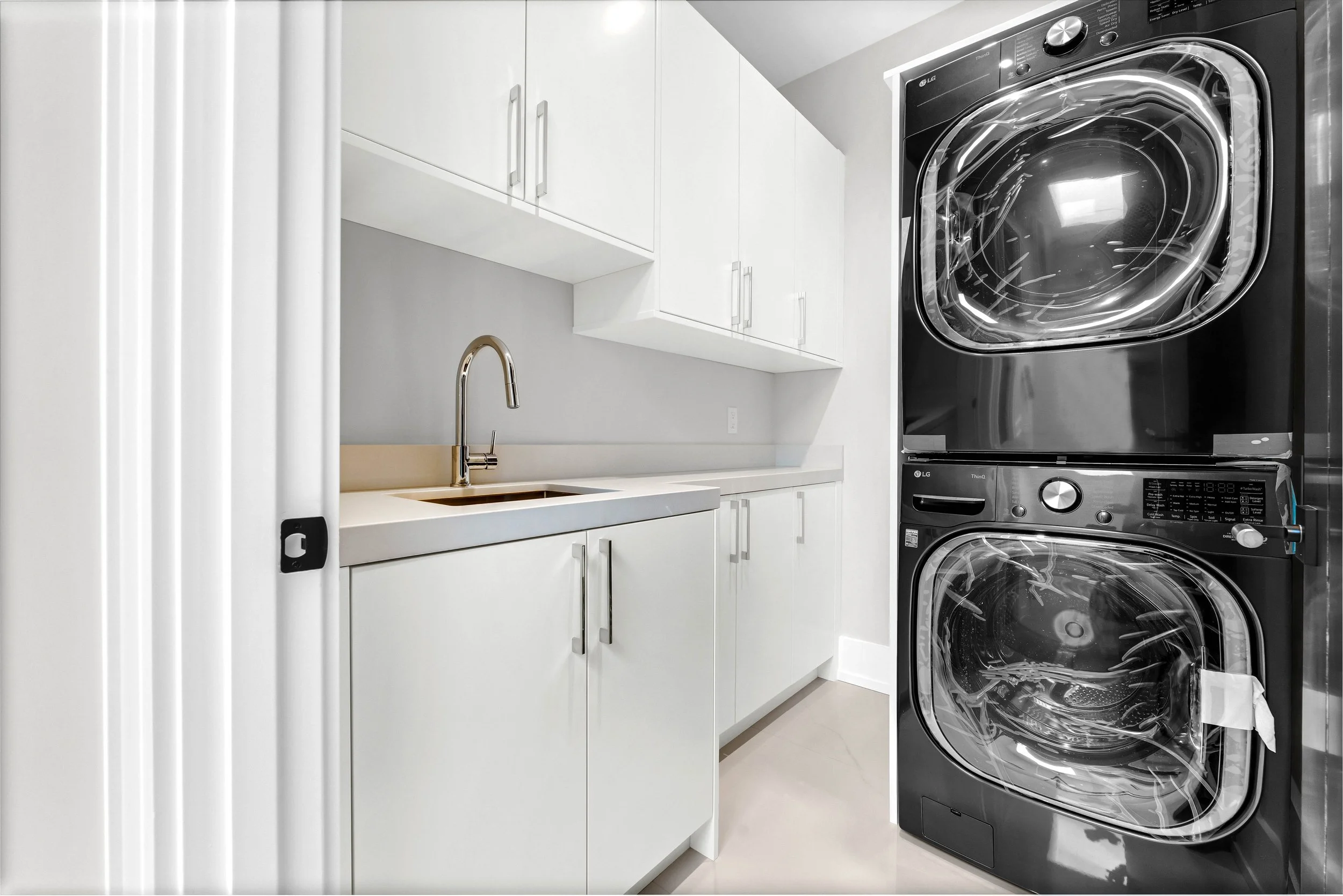 A laundry room with white cabinets, a white countertop with a sink, and a stacked washer and dryer in black.