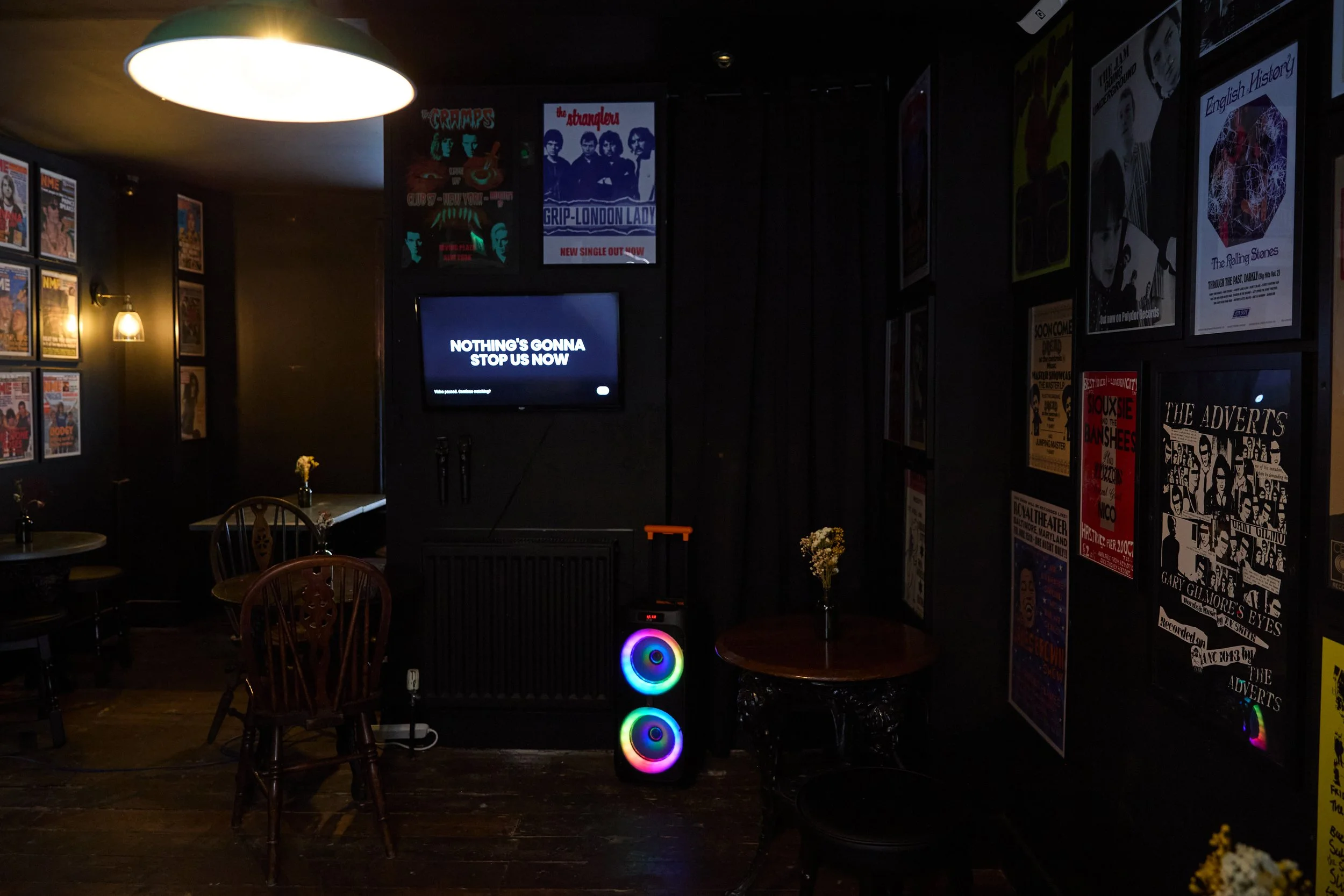 A dimly lit bar or cafe with framed posters on the walls, a small table with a flower vase, a modern speaker with colorful lights, two microphones on a stand, and a screen displaying the message "NOTHING'S GONNA STOP US NOW."