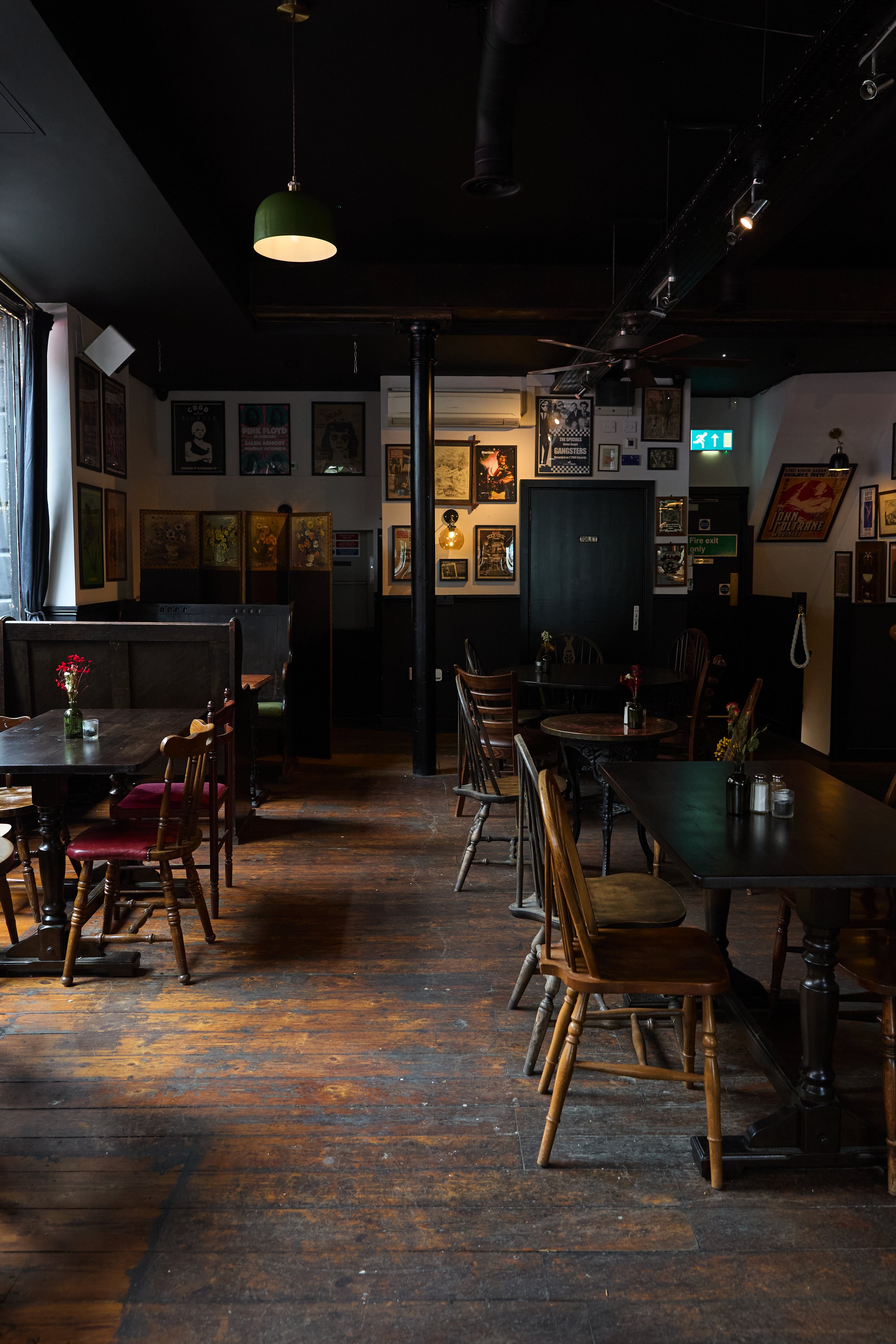Interior of a cozy, dimly lit restaurant with wooden floors and vintage decor, featuring wooden tables and chairs, flower vases, and framed art on the walls.