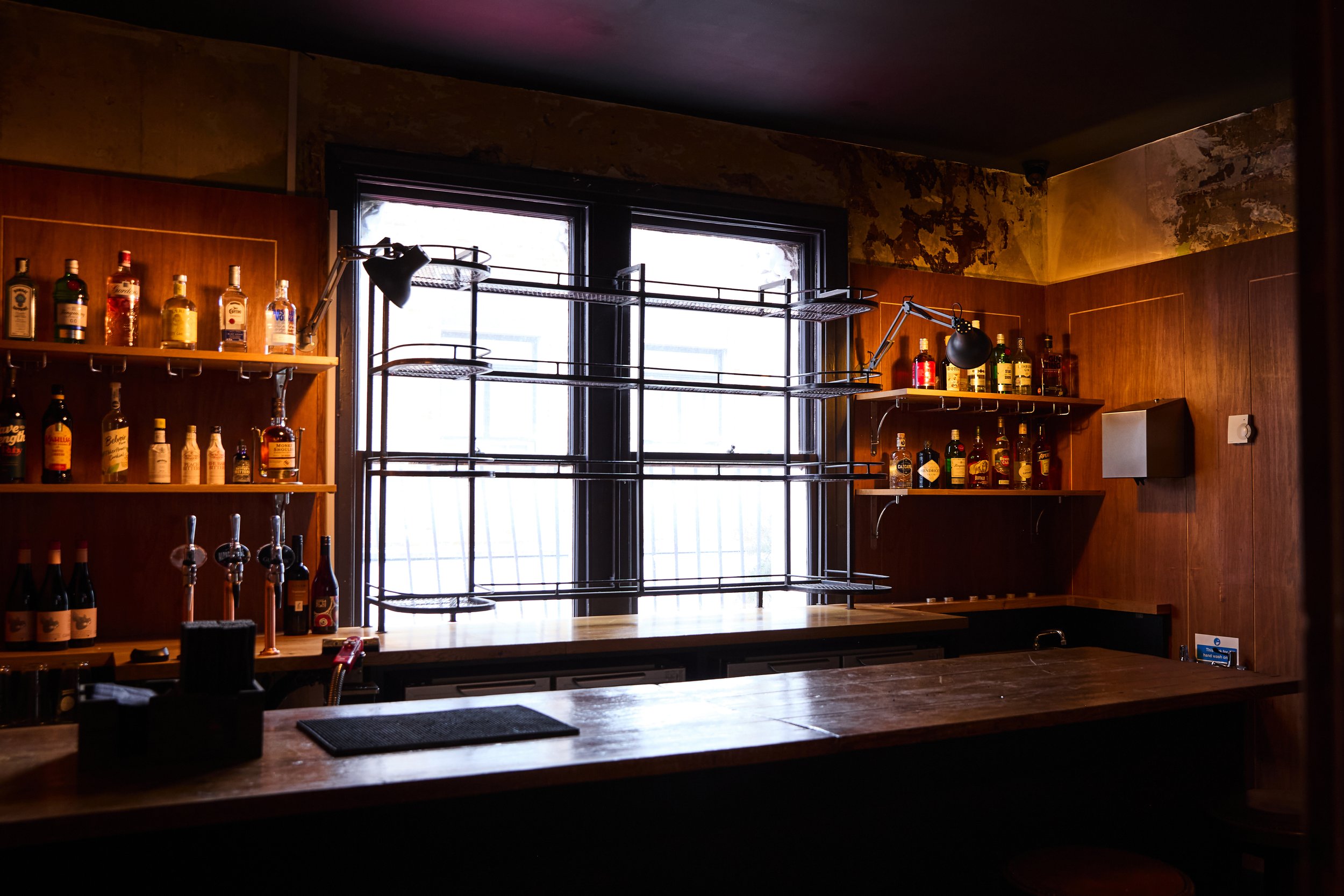 Interior of a bar with wooden shelves of liquor bottles, a window with metal bars, and a wooden counter with bar equipment.