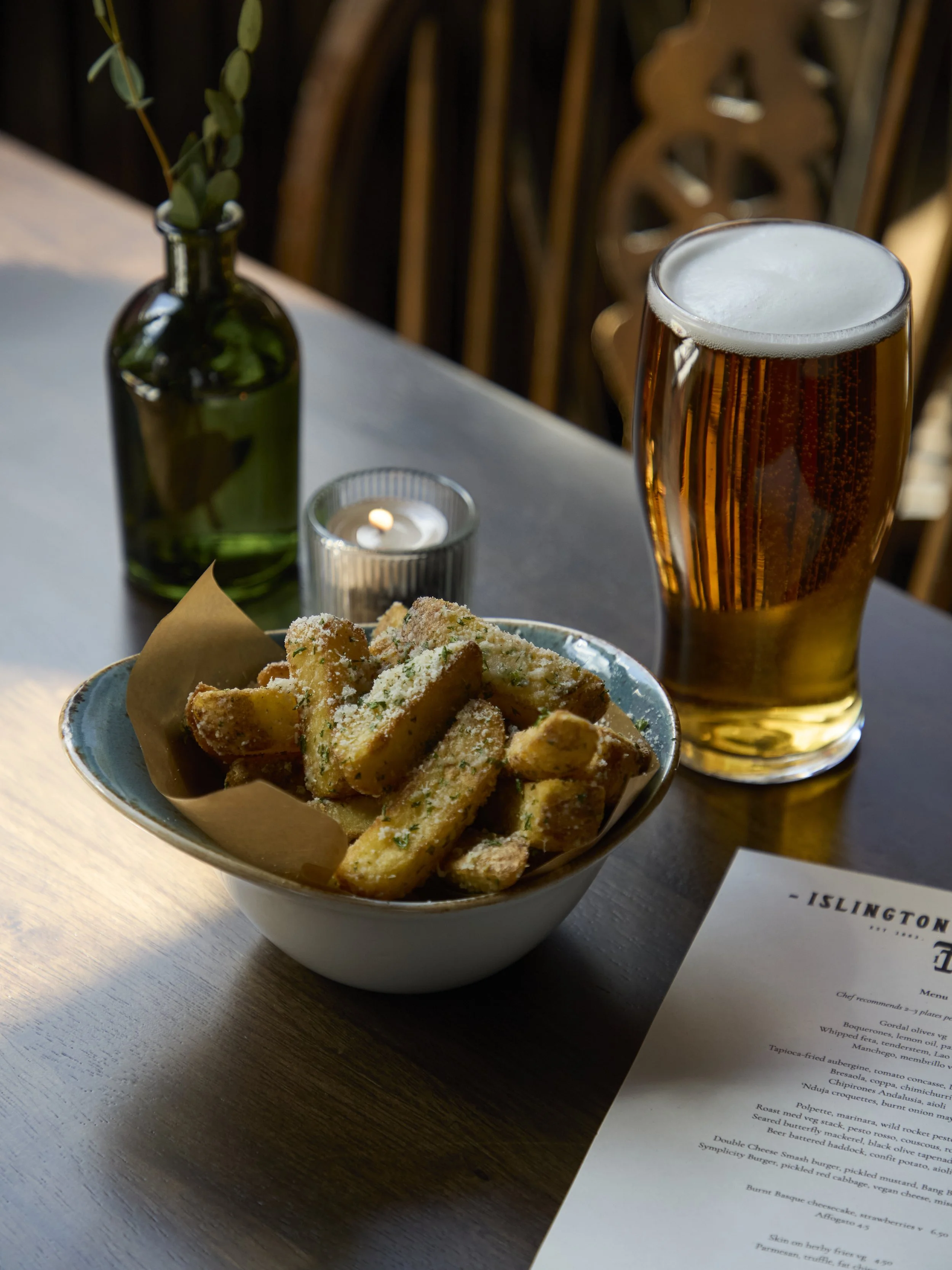 A bowl of breaded fried zucchini sticks topped with grated cheese on a table with a glass of beer, a small candle, a green vase with a plant, and a menu.