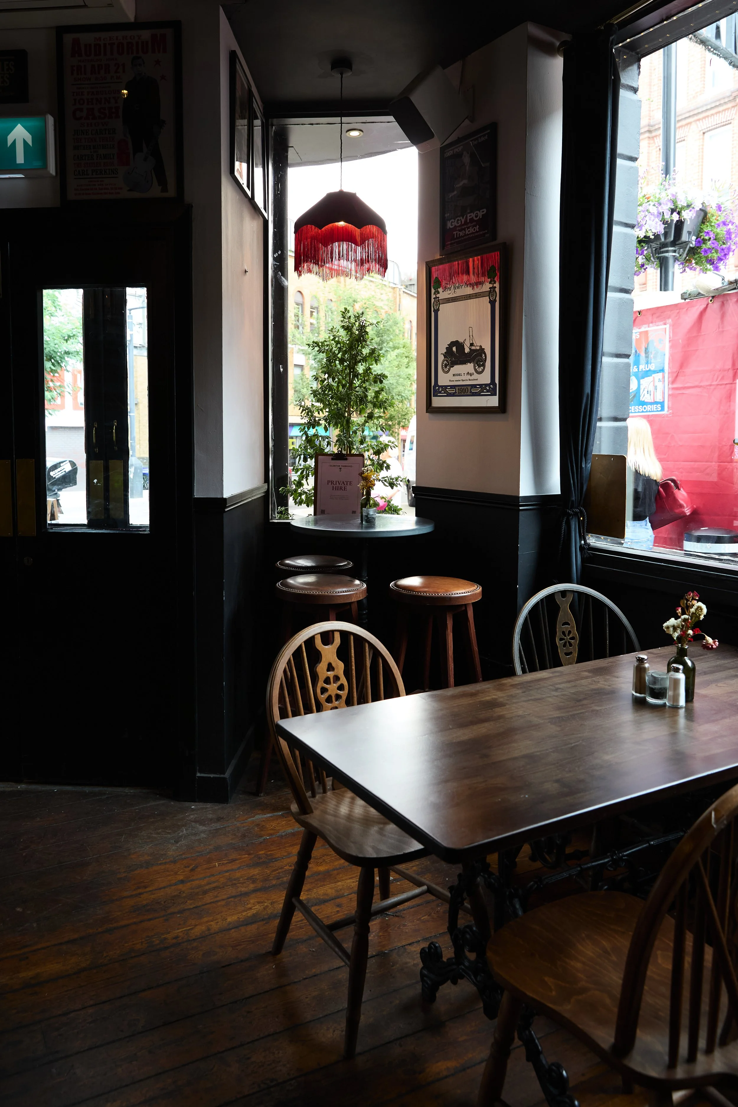 Interior of a cozy cafe with wooden tables and chairs, a small round table with bar stools near a window, decorative wall art, potted plants, and a red lampshade, with sunlight streaming in from outside.